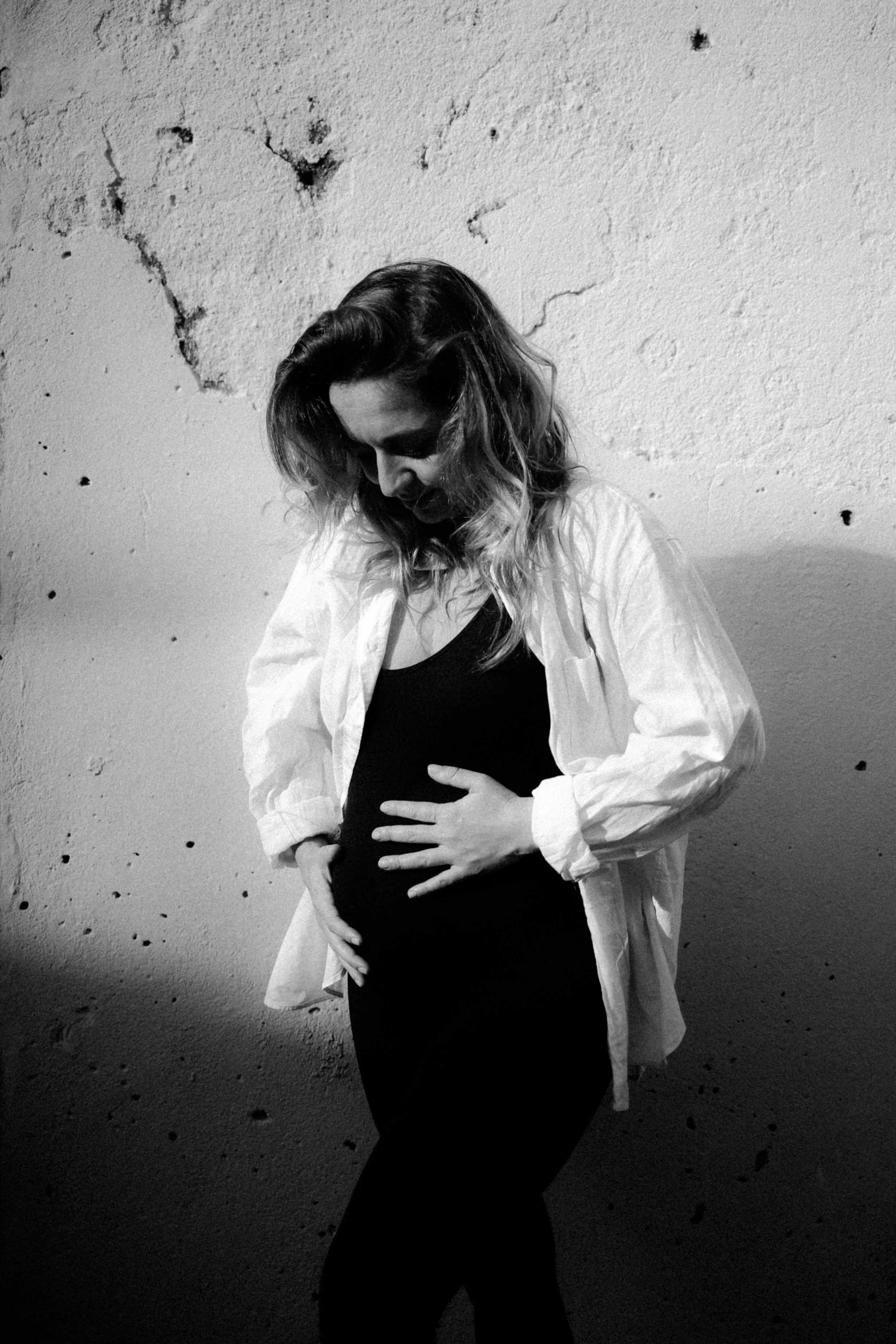 Une femme enceinte avec une main sur son ventre, vêtue d'une robe noire et d'une chemise blanche, se tient contre un mur abîmé en arrière-plan.