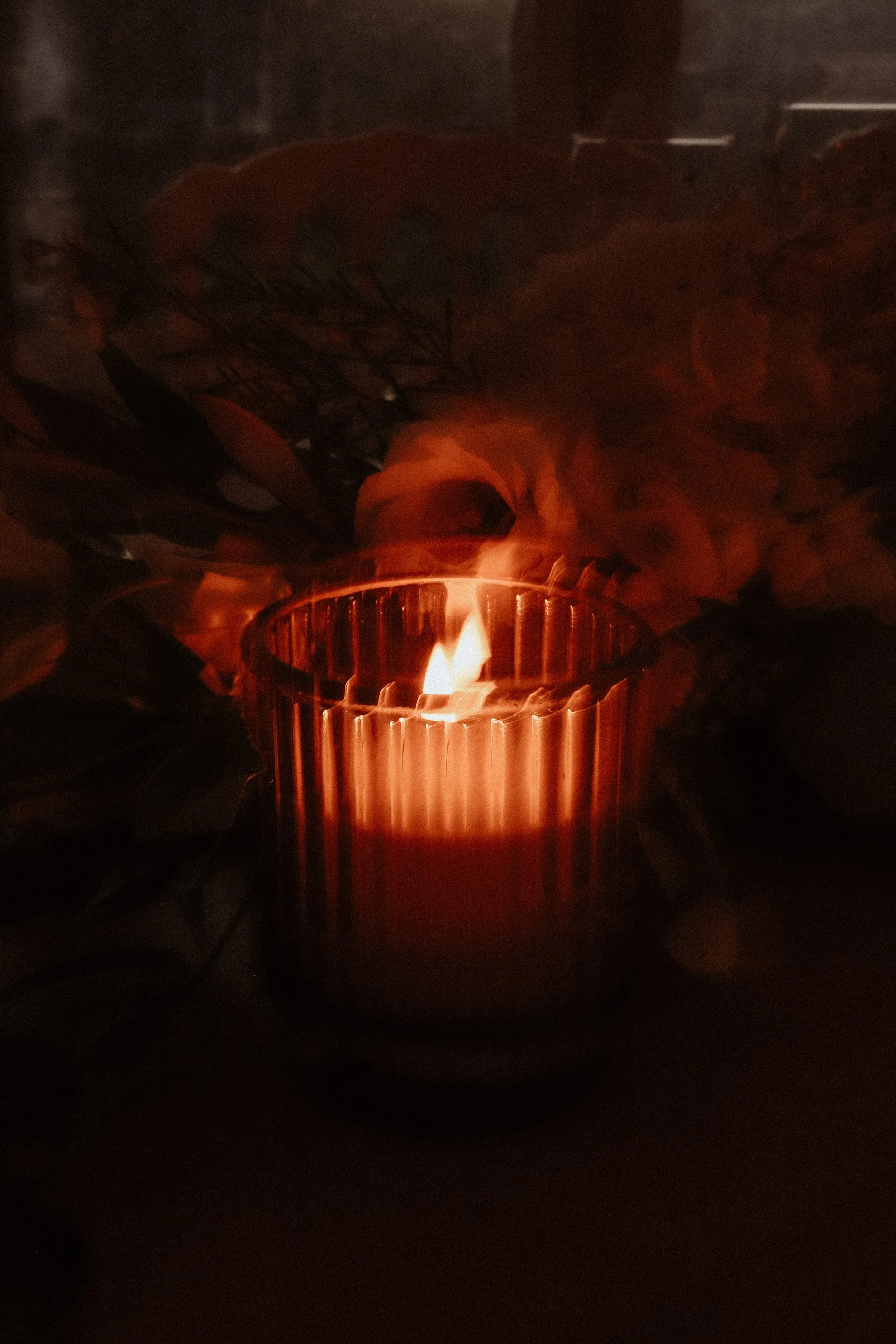 Une bougie allumée dans un récipient en verre avec des fleurs et des feuilles en arrière-plan.