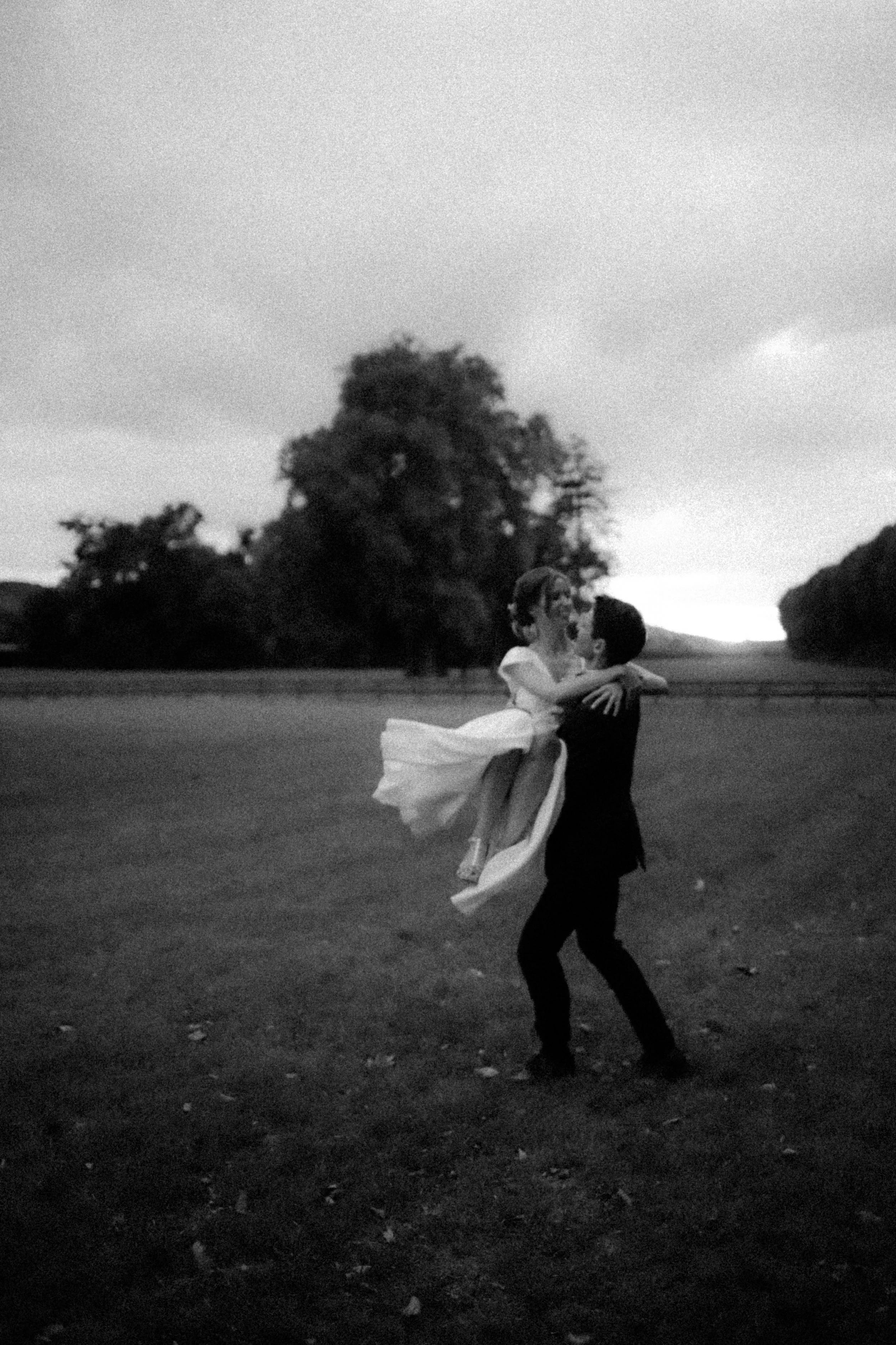 Un homme portant une femme en robe de mariage dans un champ, en noir et blanc, avec des arbres en arrière-plan.