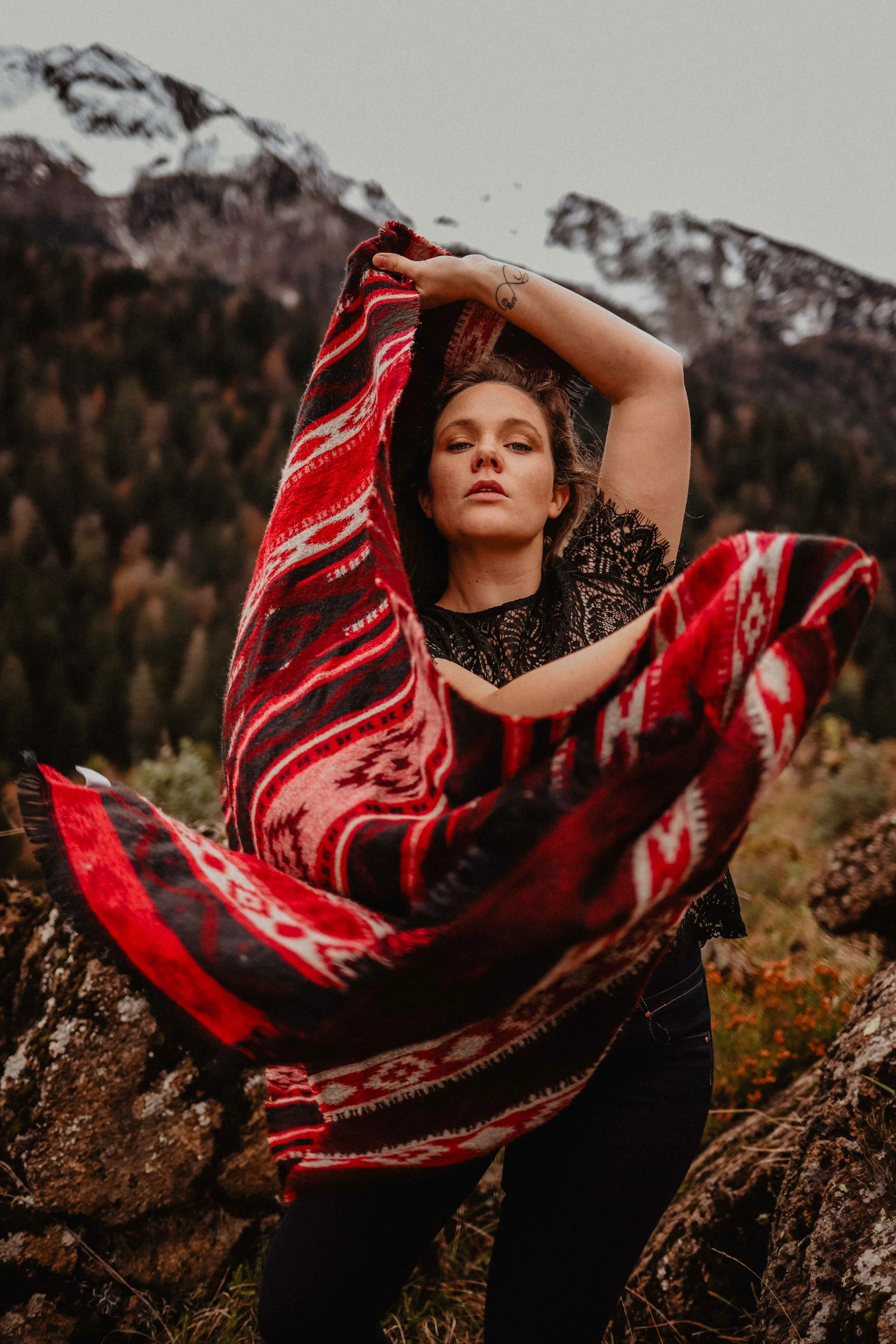 Femme dans un paysage de montagne, vêtue d'un haut en dentelle noire et tenant un tissu rouge et noir avec des motifs. Les montagnes en arrière-plan sont enneigées.