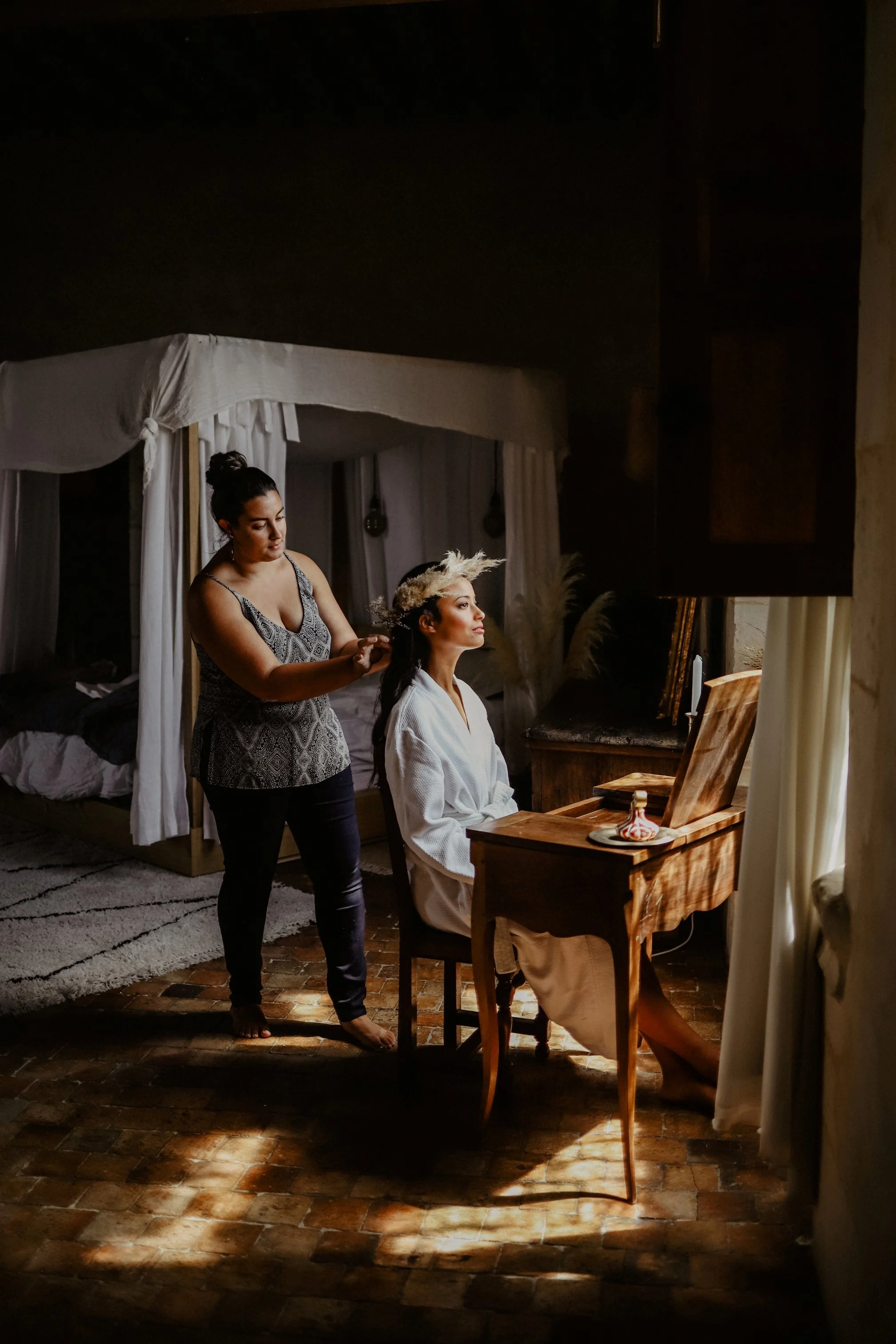 Une femme habillée d'une robe blanche assise à un bureau en bois, en train de se faire coiffer par une autre femme dans une chambre avec éclairage tamisé.
