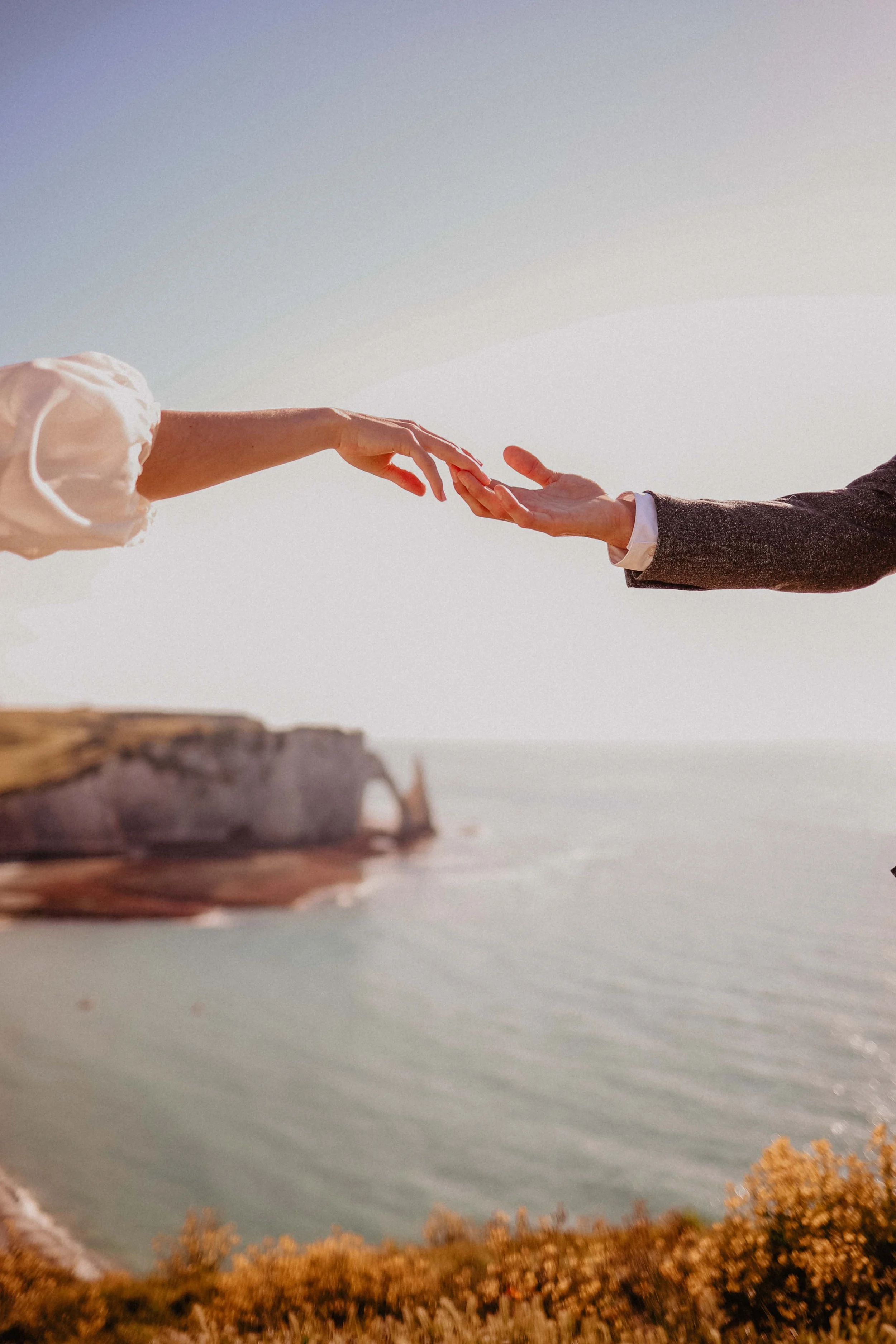 Deux personnes, apparemment un homme en costume et une femme en robe, touchent leurs mains l'une vers l'autre dans un paysage côtier au coucher du soleil.