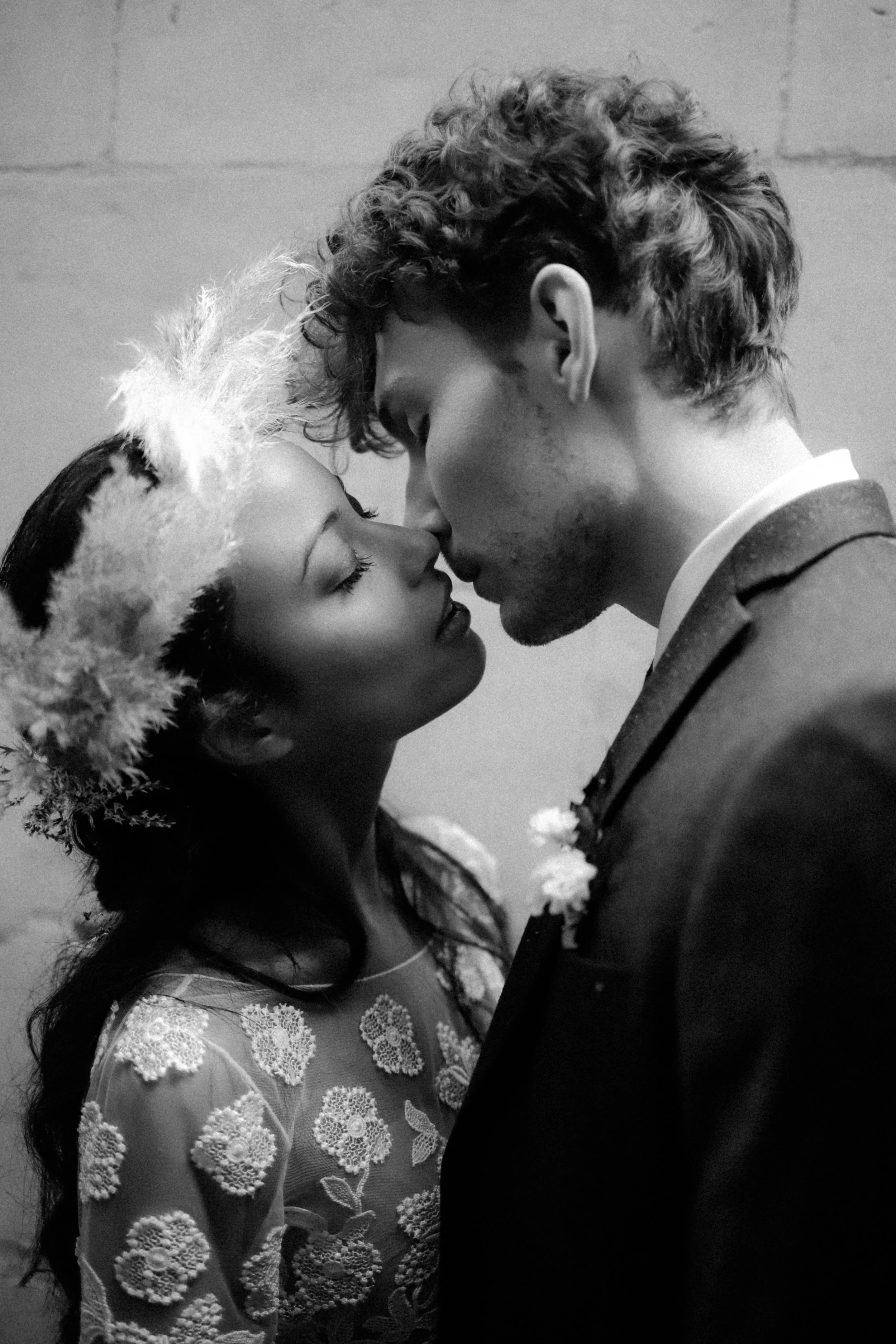 Un couple, un homme et une femme, se tenant très proches, la tête presque touchant, en un moment romantique. La femme porte une couronne en plumes et une robe en dentelle, tandis que l'homme porte un costume avec une épingle de boutonnière. L'image e
