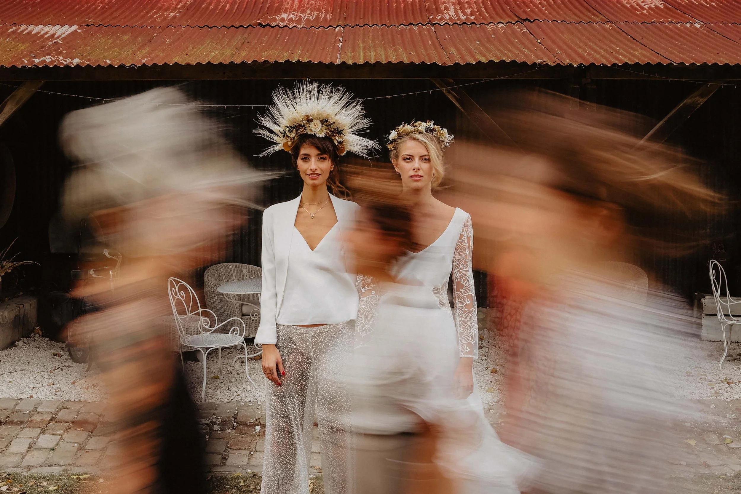 Deux femmes portant des robes blanches et des couronnes de fleurs posent devant une masonry mur avec un toit en tôle rouillée, entourées de personnes floues en mouvement.