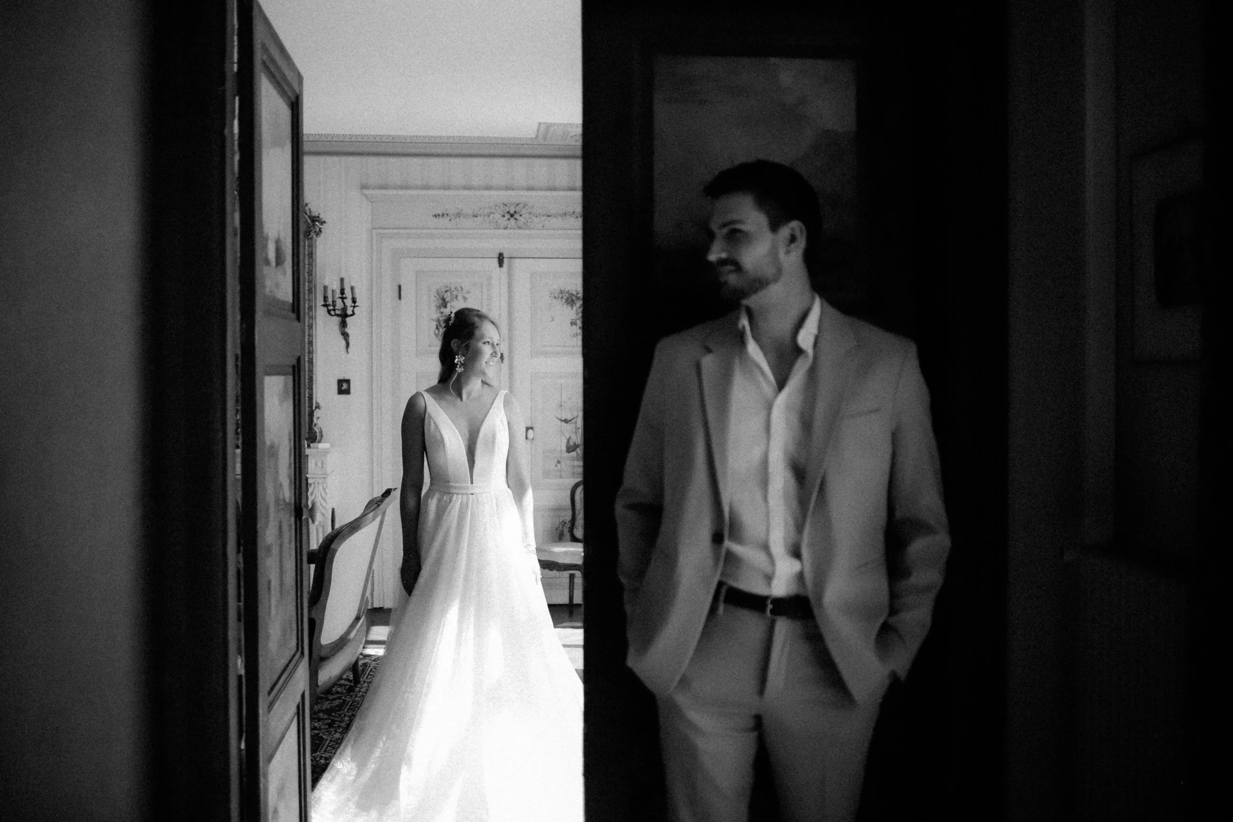 Un homme en costume debout dans une pièce sombre regardant vers une femme en robe de mariage lumineuse, dans une pièce élégante.