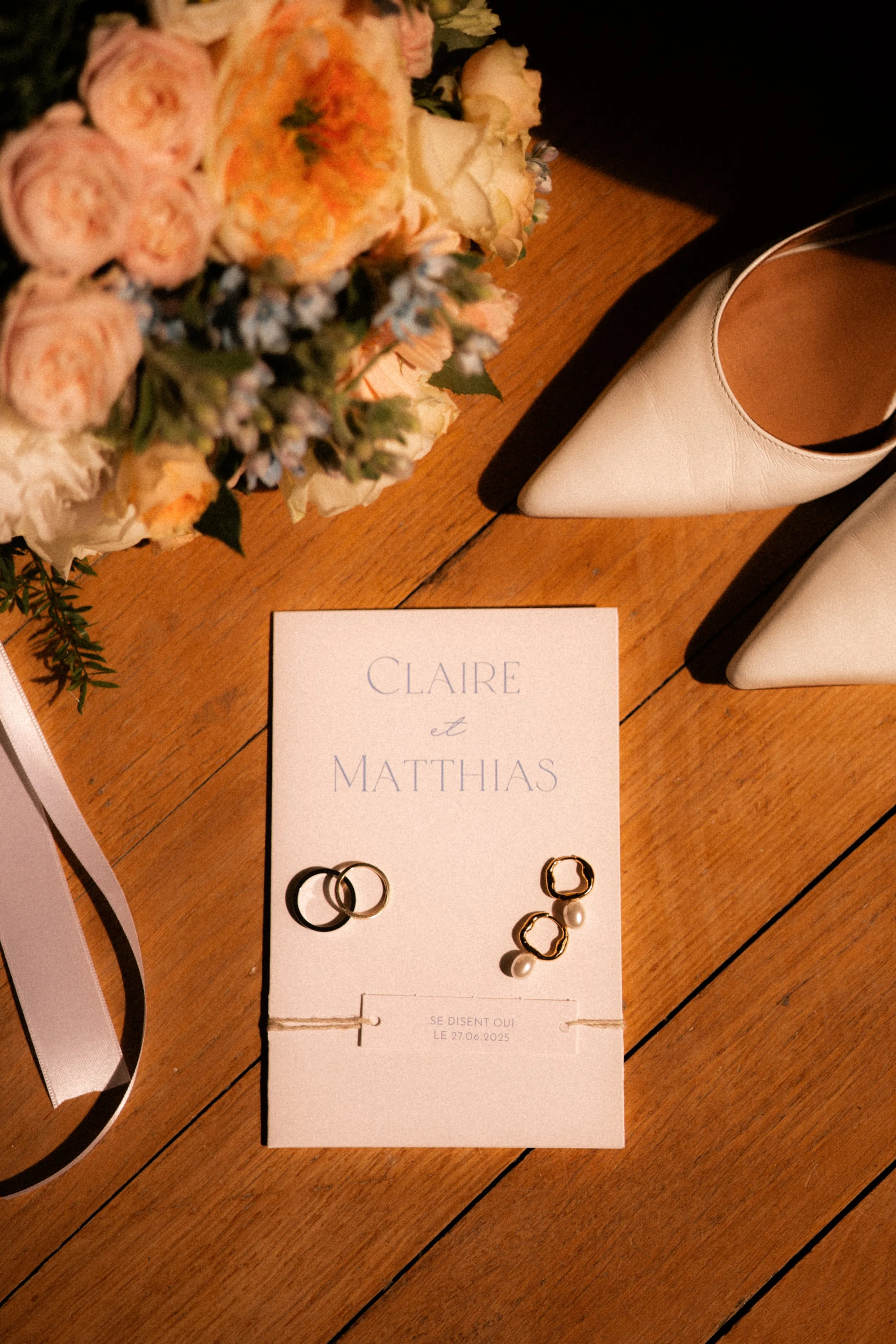 Carton d'invitation de mariage avec deux paires de bagues et des boucles d'oreilles, entouré de chaussures blanches et d'un bouquet de fleurs colorées sur une surface en bois.