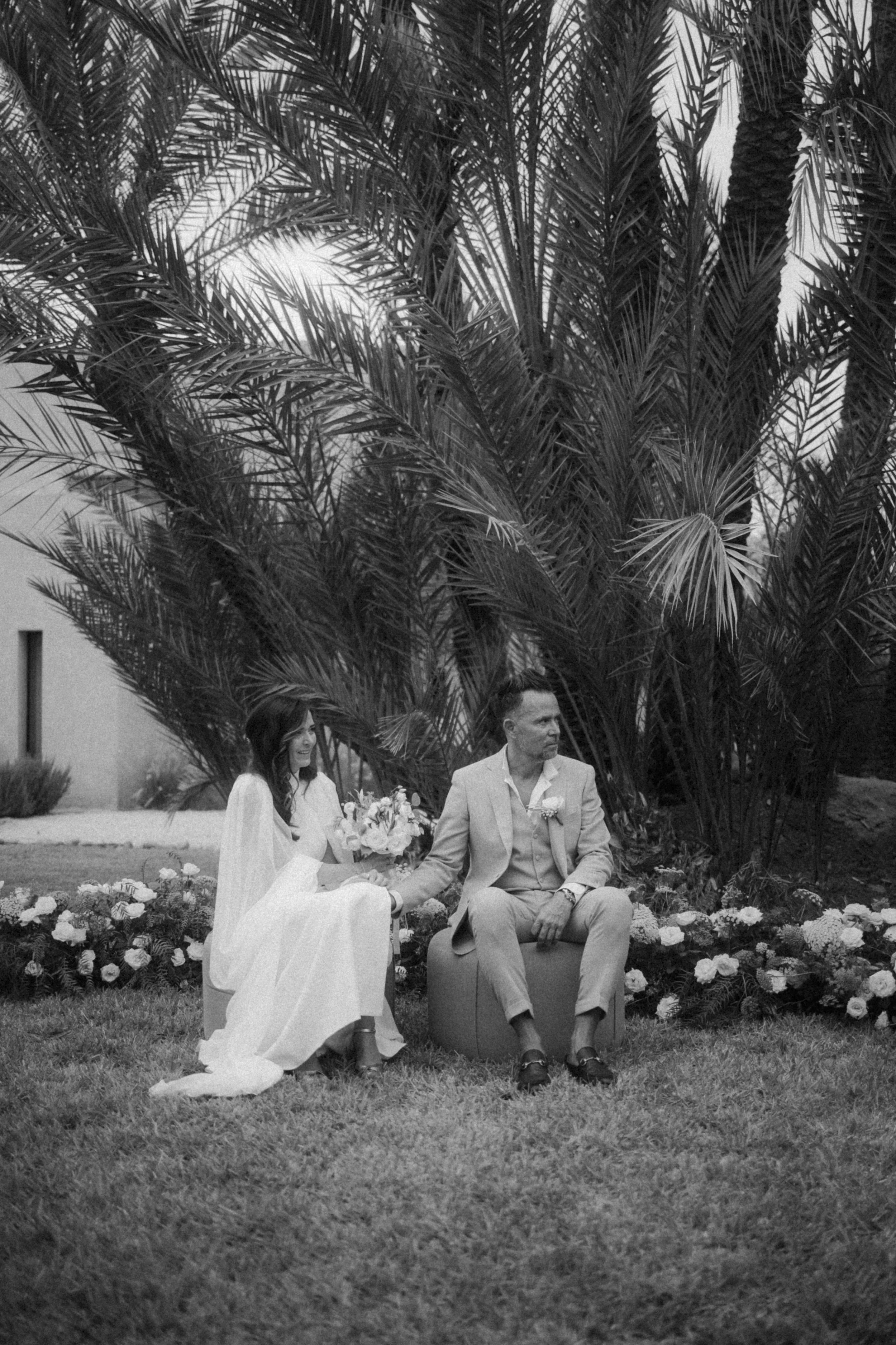 Un couple lors d'un mariage, assis sur un banc dans un jardin avec des plantes Tropicales et des fleurs en arrière-plan.