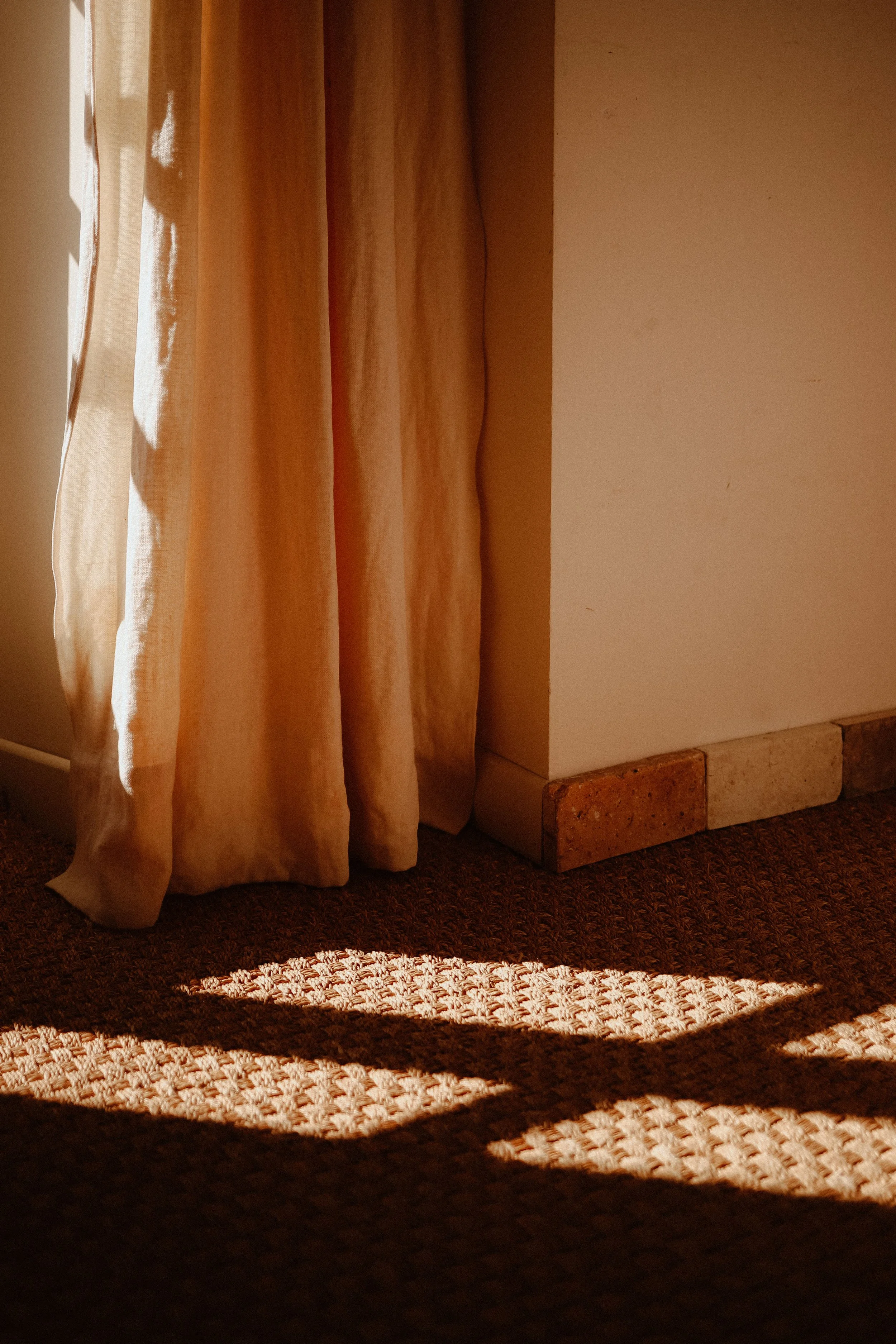 Fenêtre avec rideau beige laissant passer la lumière du soleil, créant un motif d'ombres sur le tapis.