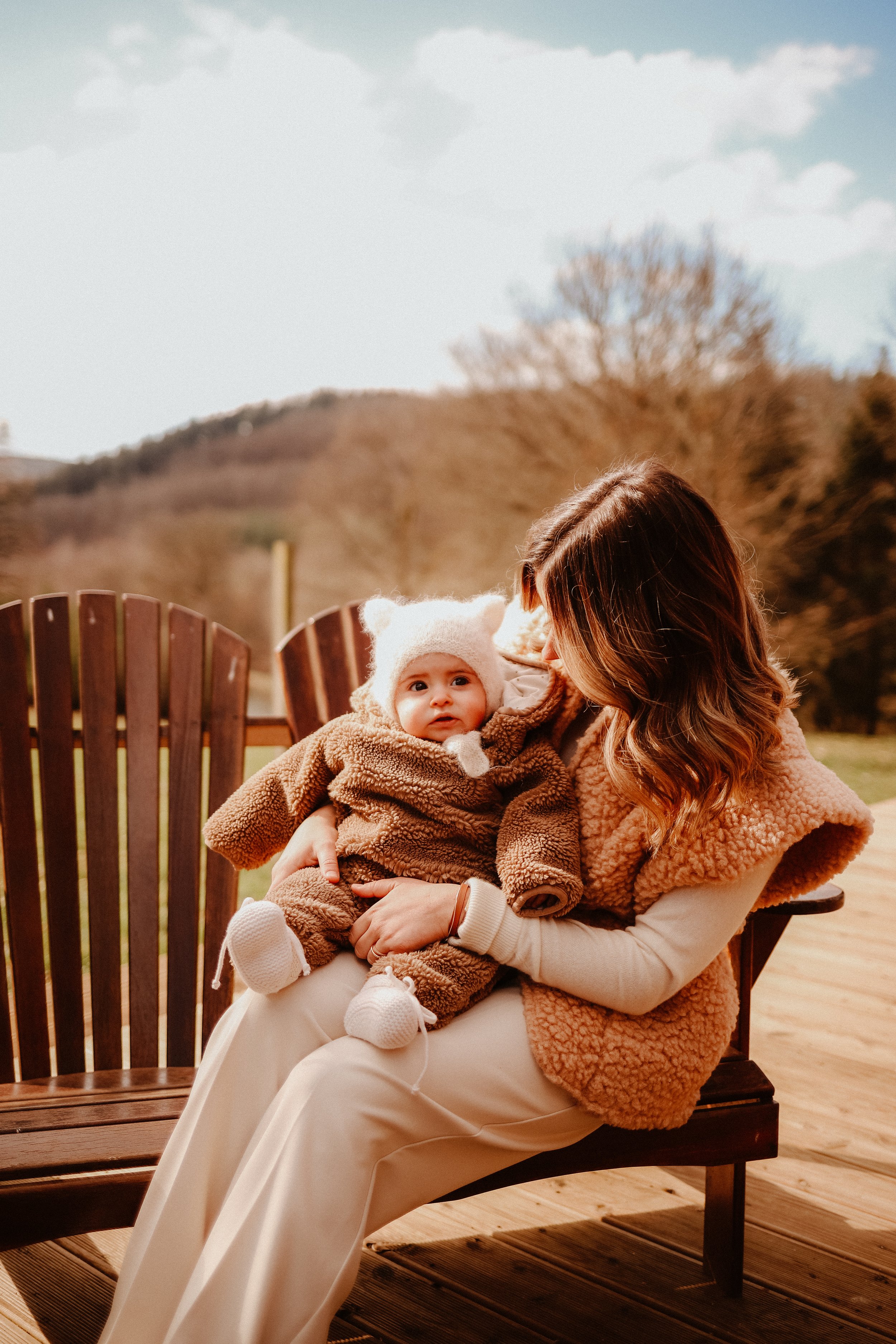 Une femme tenant un bébé assis sur un banc en bois à l'extérieur, avec des montagnes et des arbres en arrière-plan, par une journée ensoleillée.