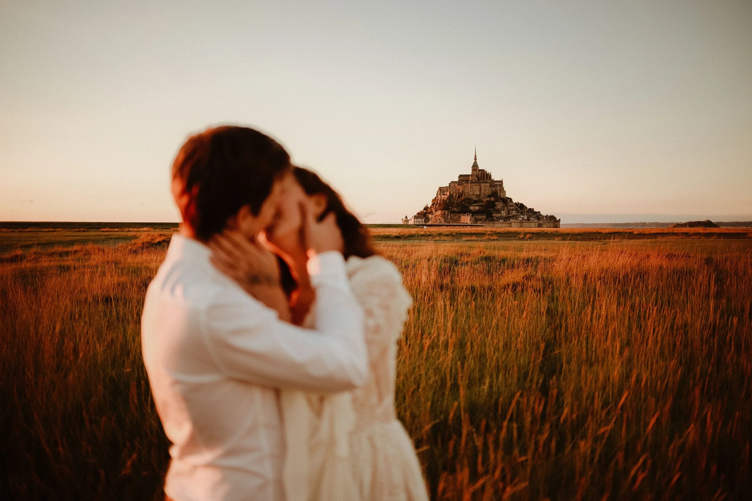 Un couple s'embrassant dans un champ avec le Mont Saint-Michel à l'horizon lors du coucher de soleil.