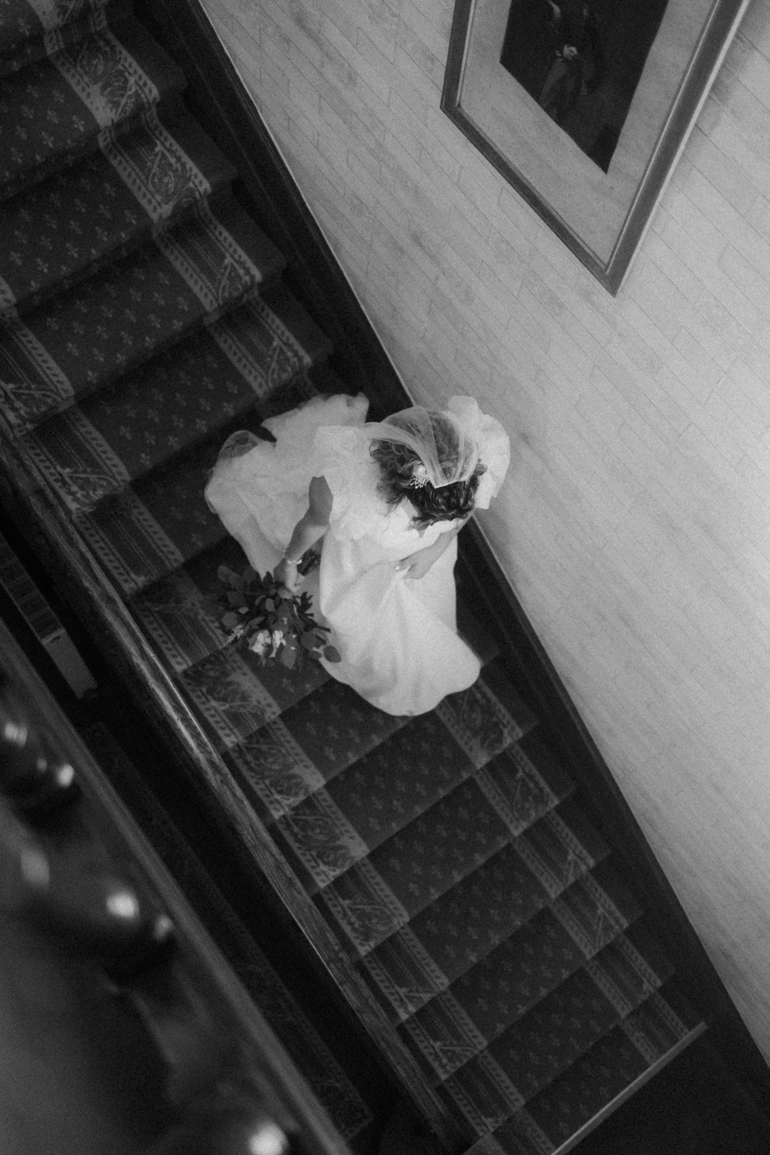 Une femme en robe de mariée descendant un escalier en chaîne, vue de dessus dans une image en noir et blanc.