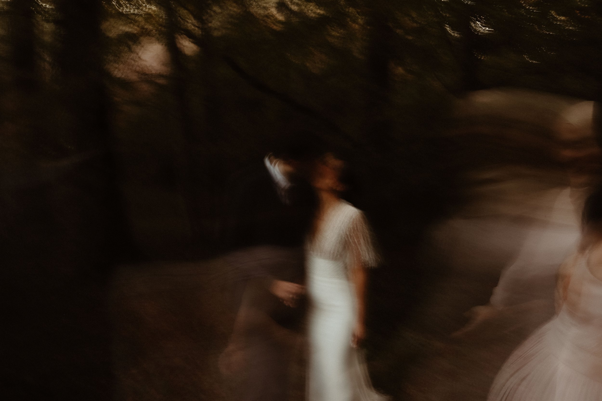 Photo floue de deux femmes en robe, une en blanc, dans un environnement sombre avec des arbres ou des branches.
