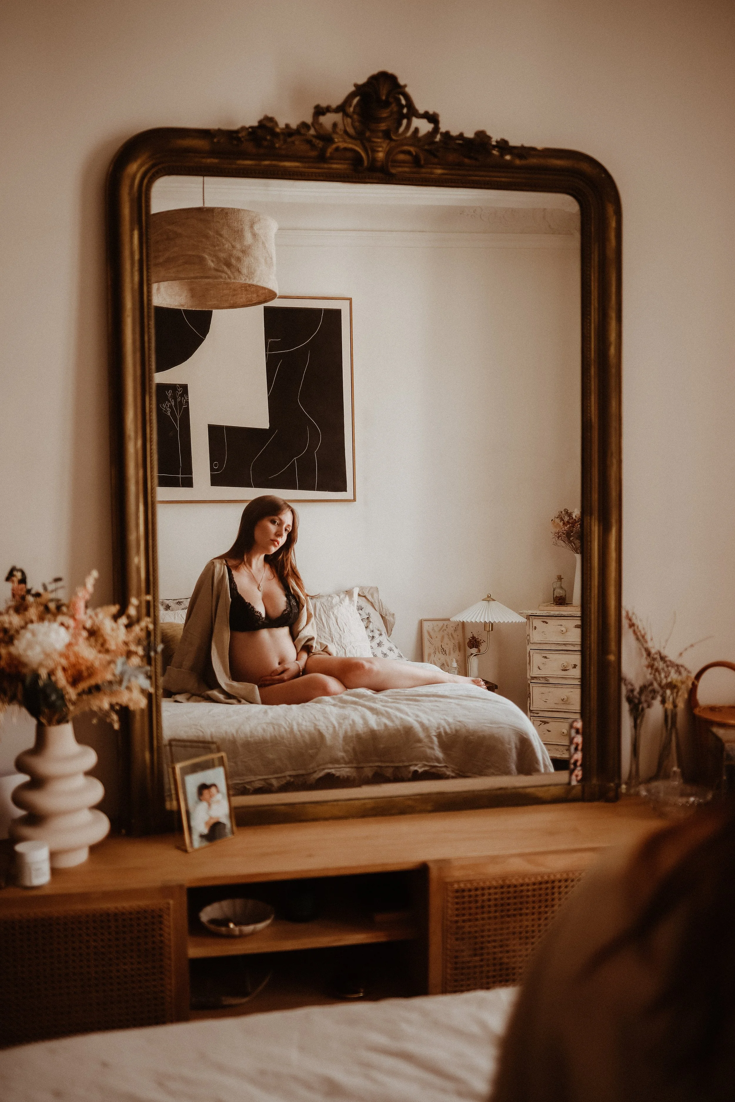 Une femme enceinte en lingerie noire et robe beige clair, assise sur un lit dans une chambre décorée de façon vintage, vue à travers un grand miroir doré.