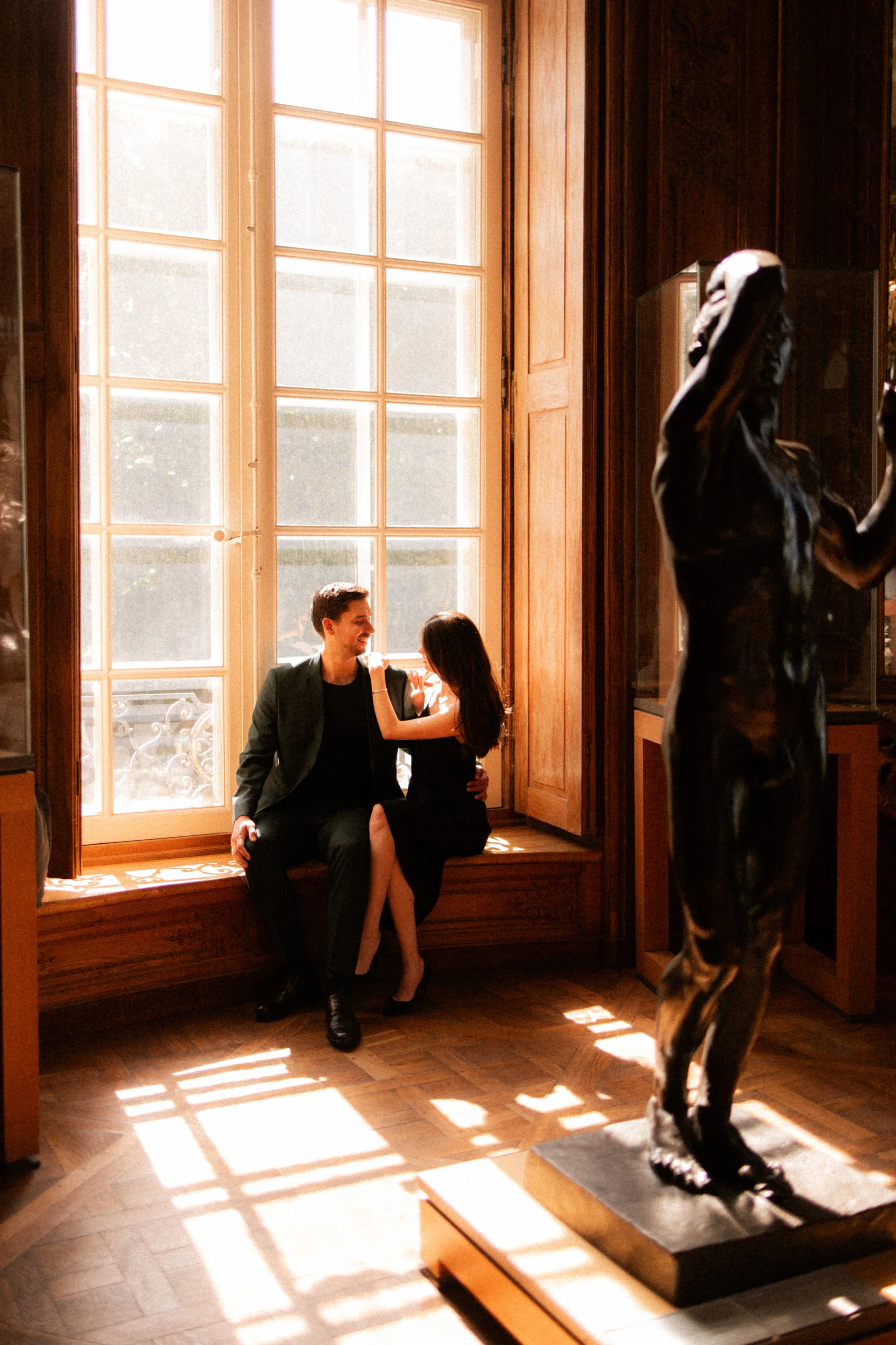 Un homme et une femme assis côte à côte près d'une grande fenêtre en bois dans une salle d'exposition, avec une sculpture en bronze d'un homme au premier plan.