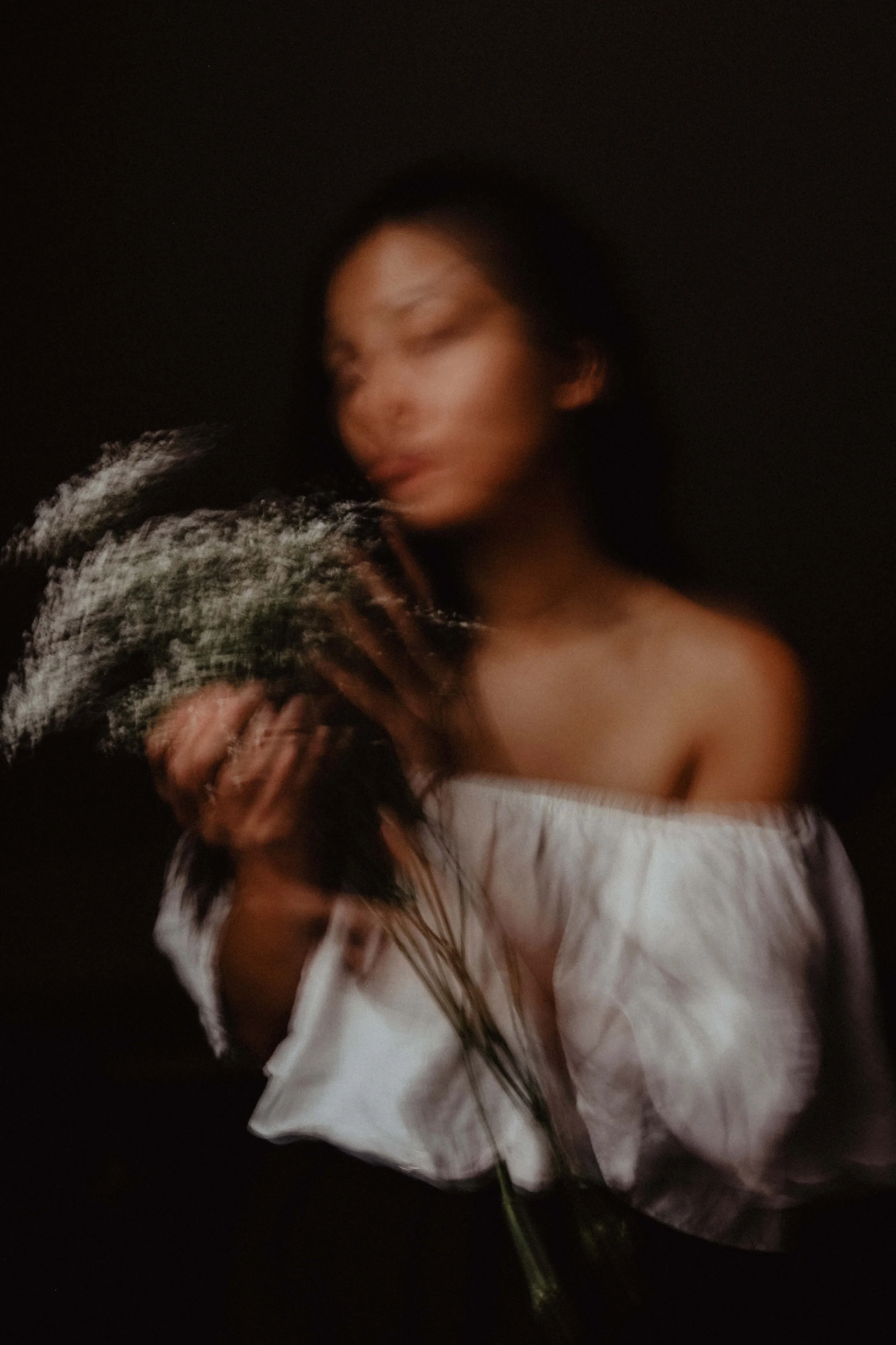 Une femme avec une peau claire et des cheveux longs tient un bouquet de fleurs. La photo est floue avec un effet de mouvement, sur un fond noir.