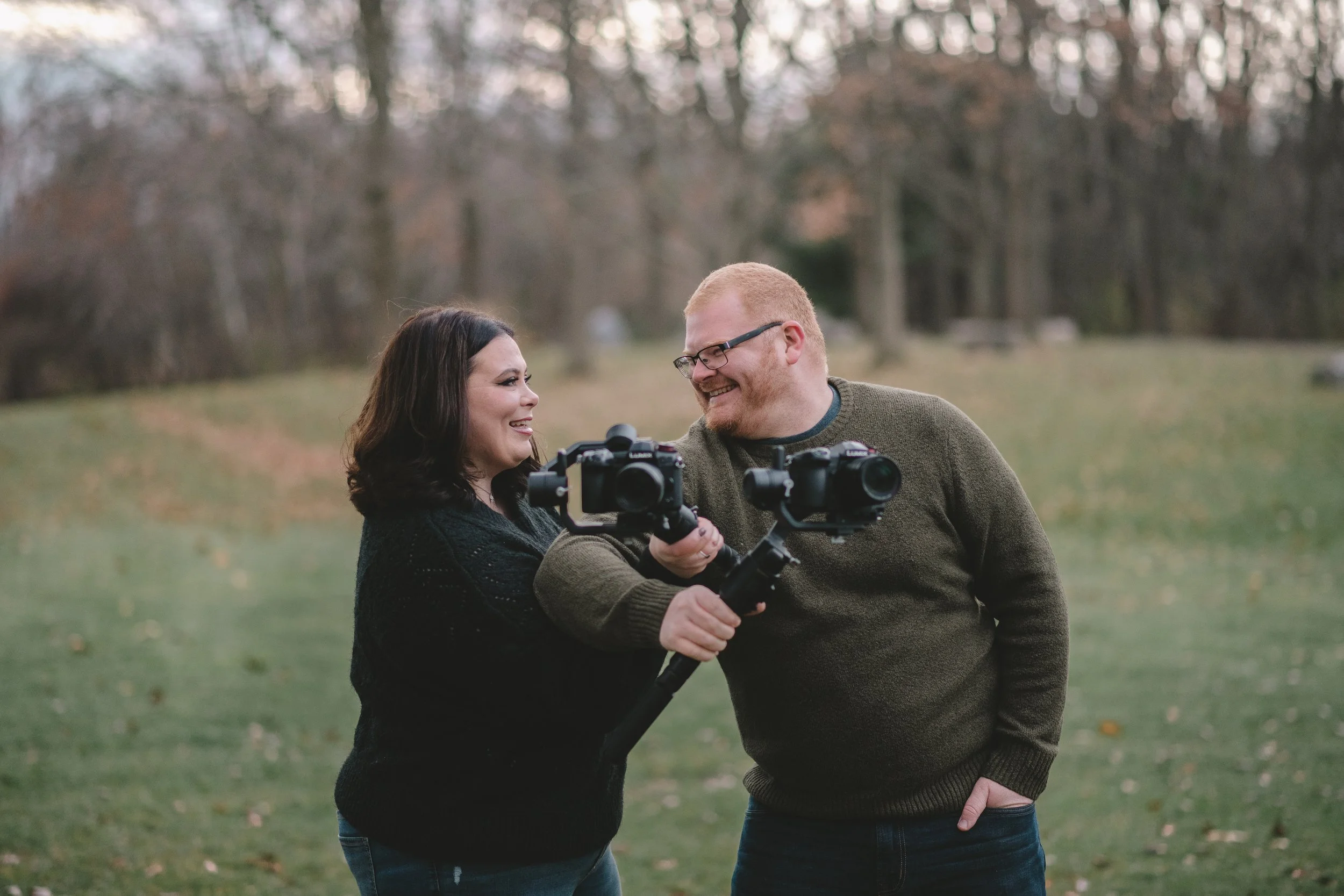 A smiling woman and man holding a camera together in a park with trees in the background.