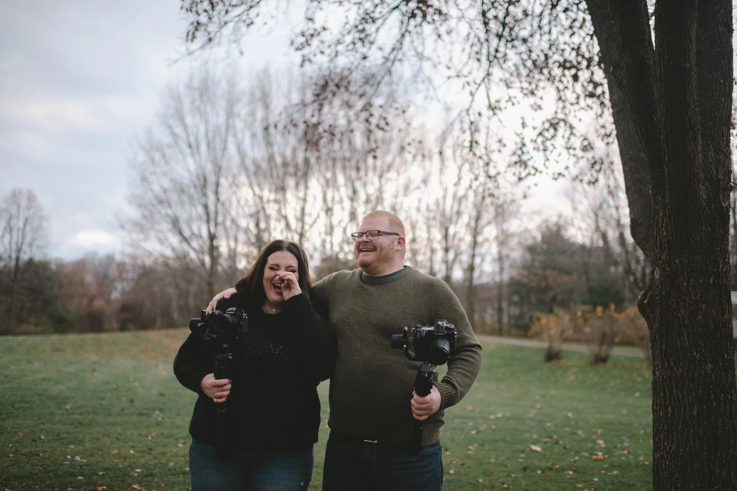 A woman and a man standing outdoors on a cloudy day, laughing and holding cameras. The woman has dark hair and is covering her nose, while the man, with light hair and glasses, has his arm around her shoulder.