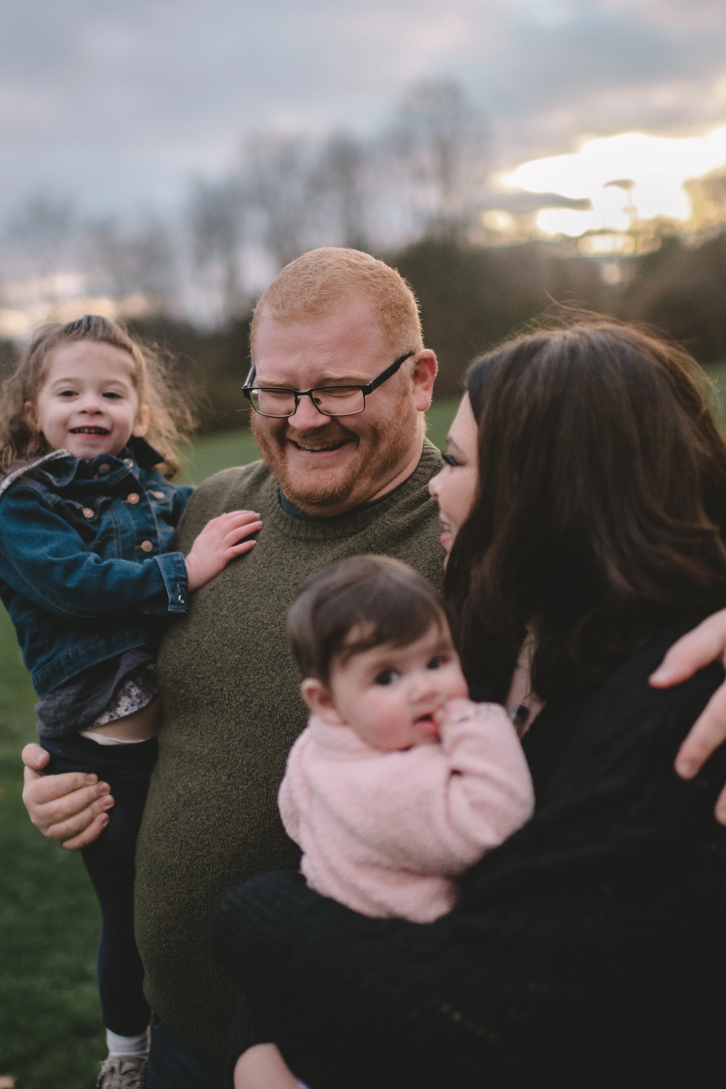 A family of four outdoors; a man with glasses, an woman, a young girl on the man's shoulder, and a baby in the woman's arms, all smiling and enjoying the moment at sunset.