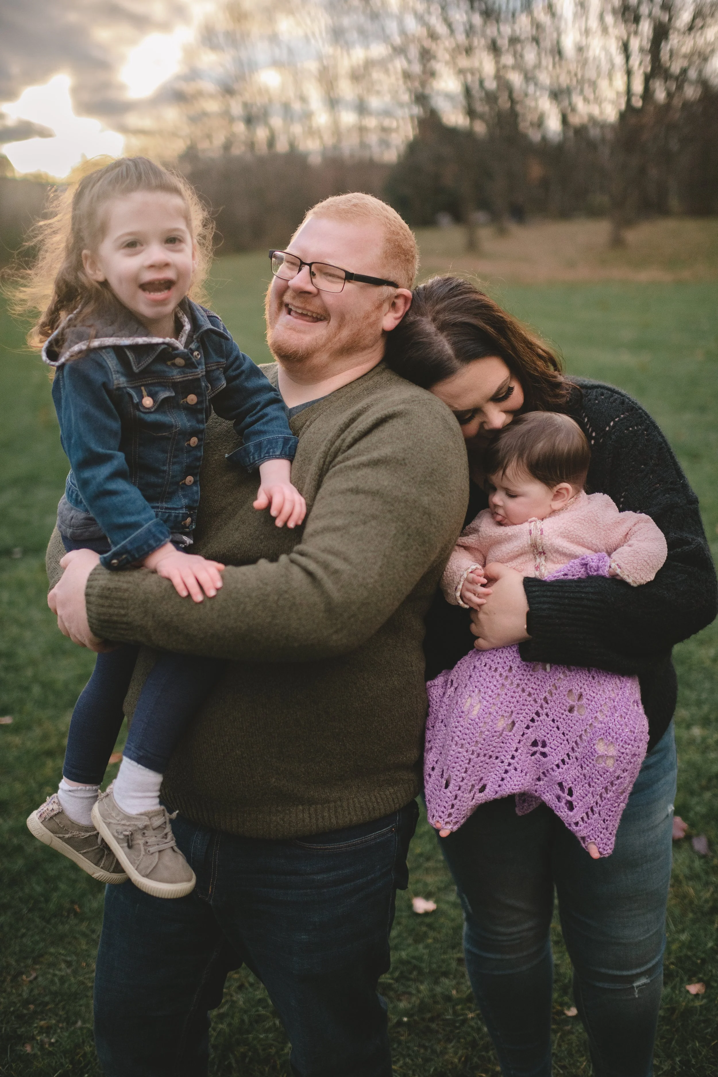 A family of four enjoying an outdoor moment. The father, with glasses and a green sweater, is holding a young girl with curly hair and a denim jacket. The mother, with dark hair and a black sweater, is holding a baby wrapped in a pink blanket with purple crochet edging. They are on a grassy field with trees in the background during sunset.