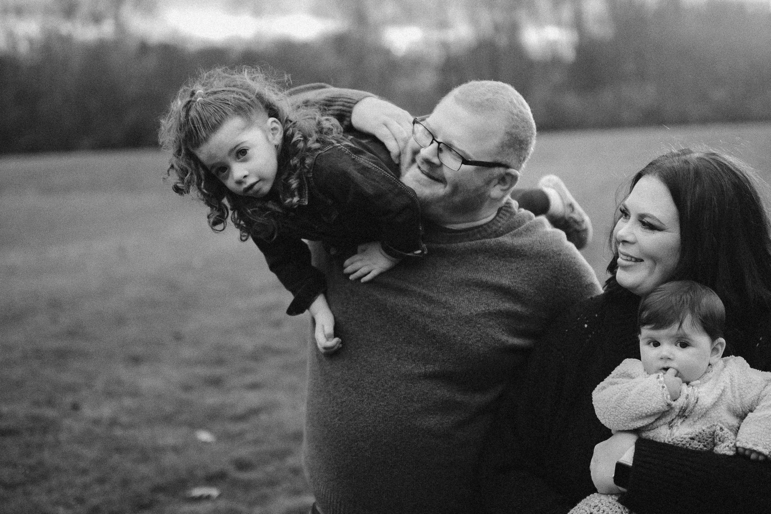 A family outdoors, with a man holding a girl on his shoulder, a woman holding a baby, all smiling, in black and white.