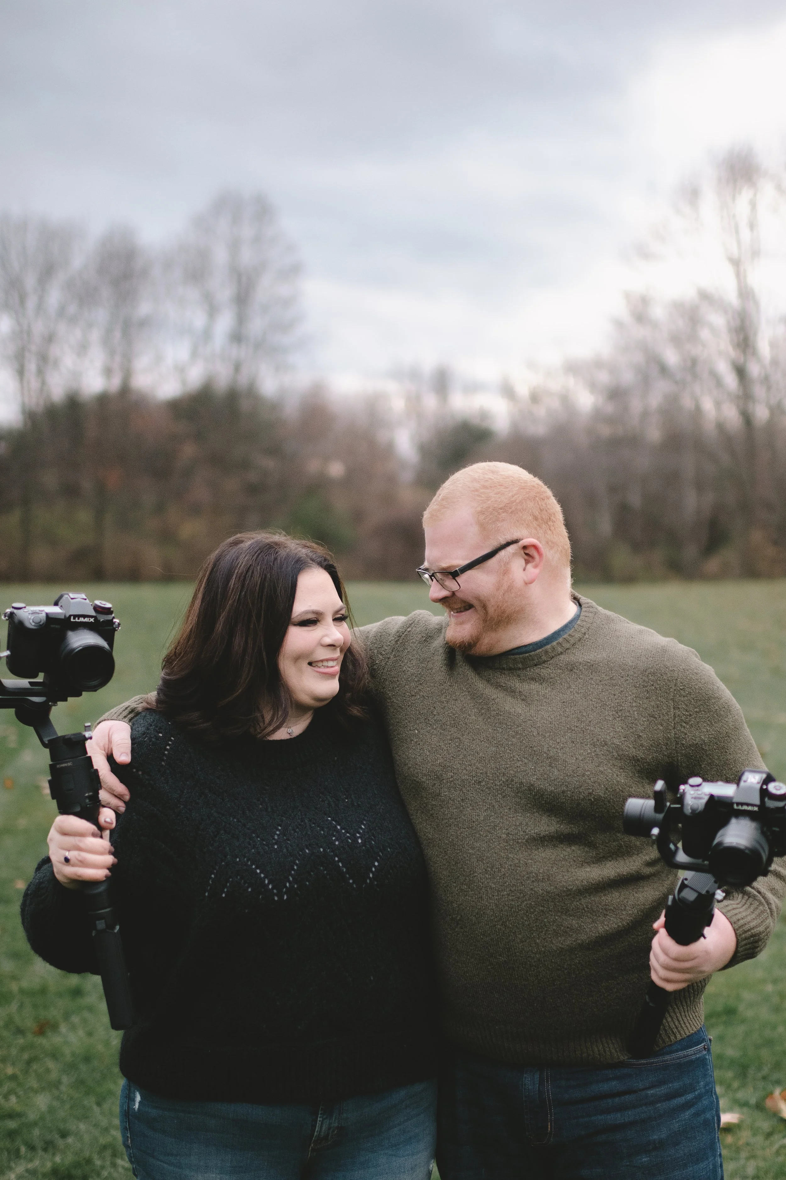 A man and woman standing close together outdoors on a cloudy day, smiling and holding cameras with stabilizers, with leafless trees and a grassy field in the background.