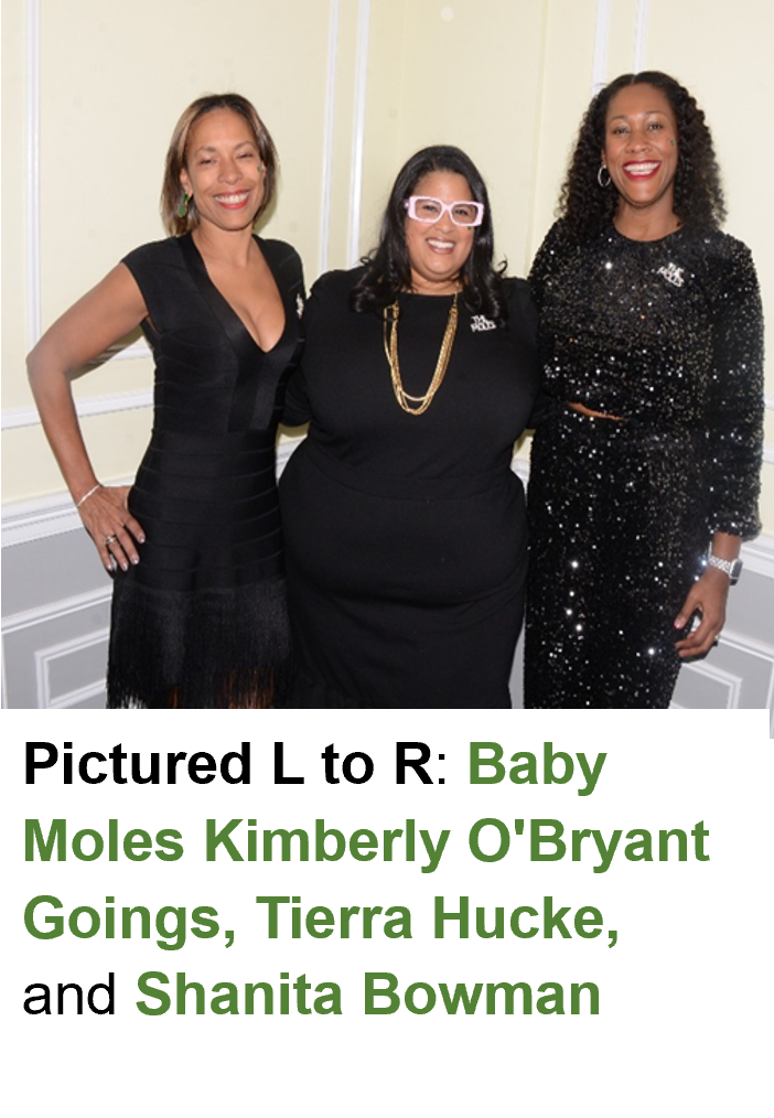 Three women standing together, smiling, dressed in black outfits, indoors with cream-colored walls.