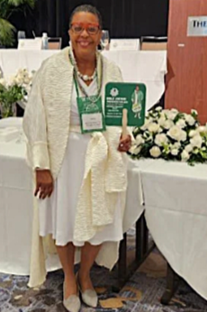 A woman standing at a conference or event, holding a green sign and smiling. She is wearing glasses, a cream-colored shawl, a white dress, and a name badge, with floral arrangements around her in an indoor setting.
