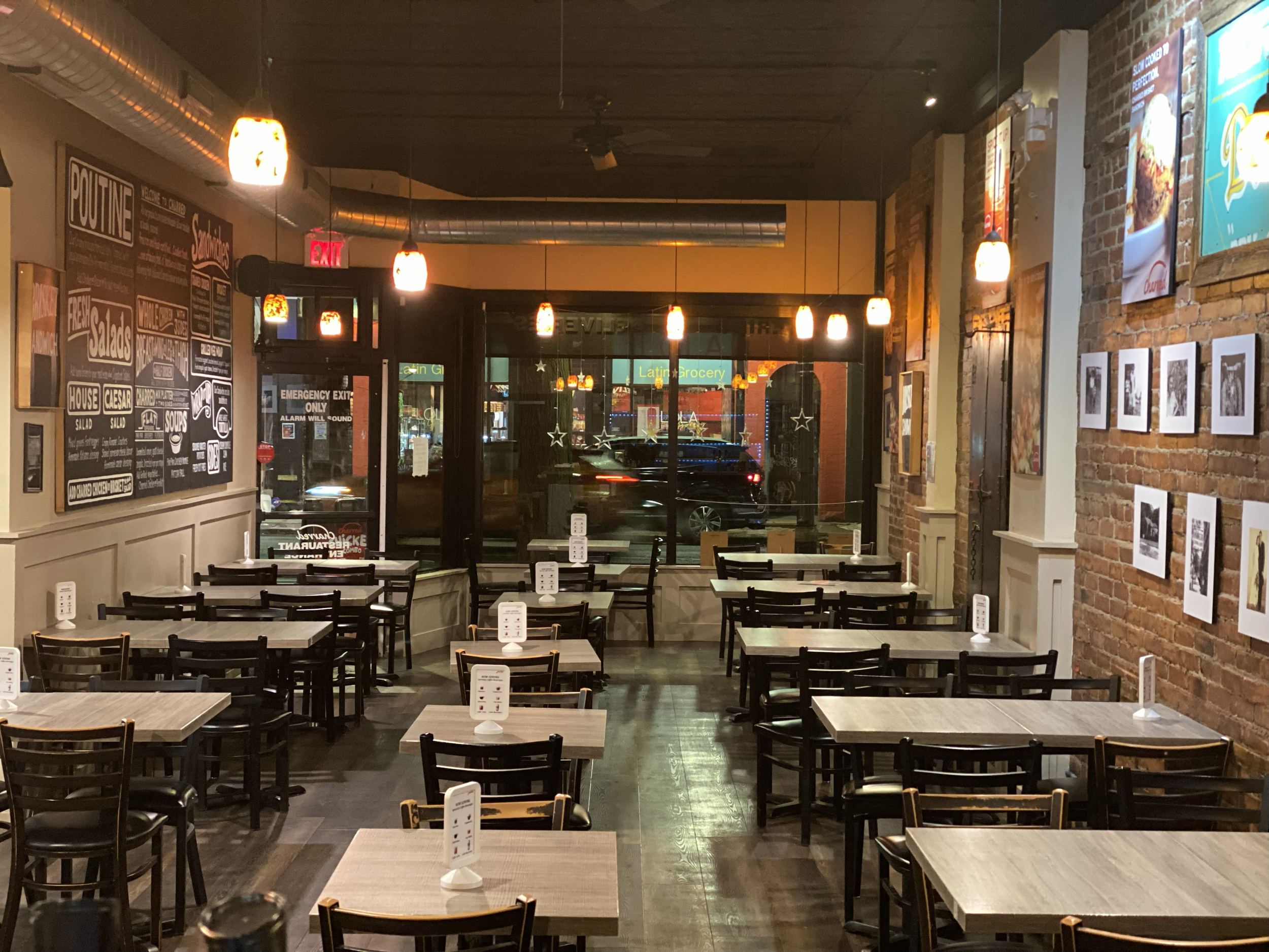 Empty restaurant with wooden tables and black chairs, brick wall with framed pictures, and a large chalkboard menu on the wall. Exterior neon signs visible through the front window.