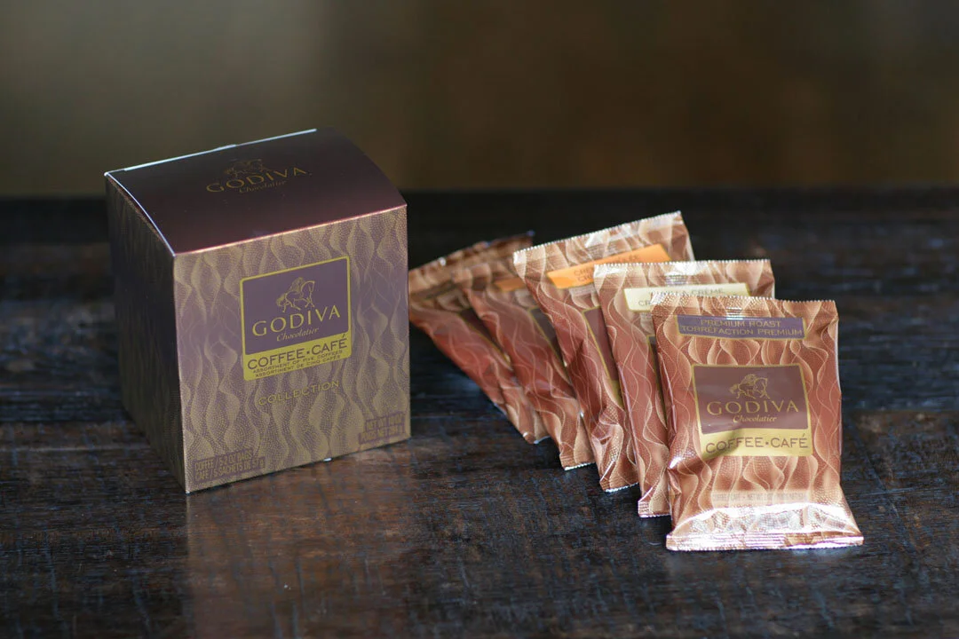 A box of Godiva coffee coffee capsules and several capsules next to it on a dark wooden surface.