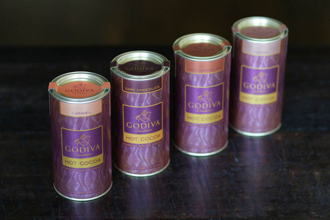 Four cans of Godiva hot cocoa in different flavors, including caramel, dark chocolate, and milk chocolate, placed on a dark wooden surface.