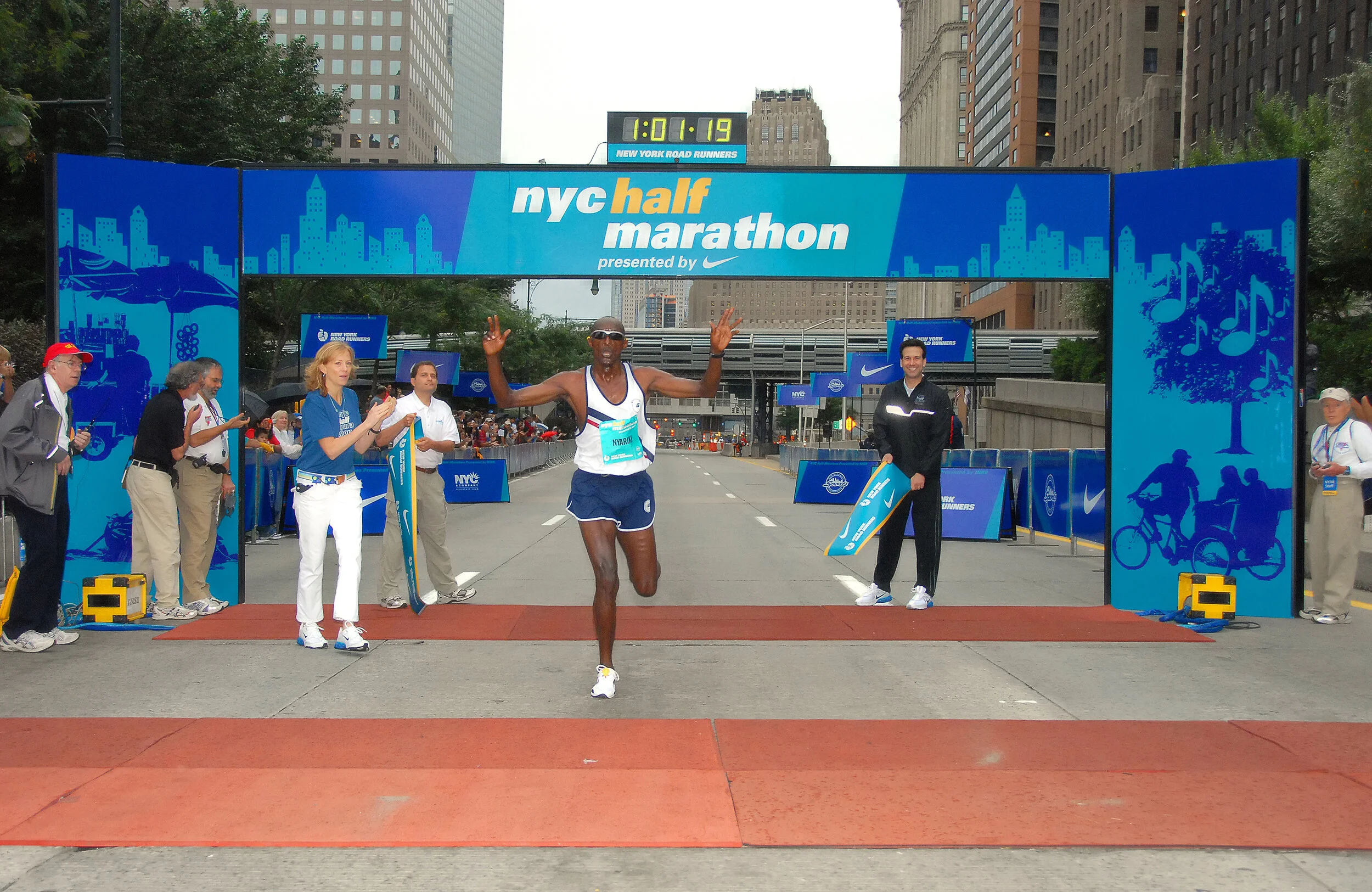 A male marathon runner crosses the finish line at the NYC Half Marathon, with people cheering on the sides and a cityscape in the background.