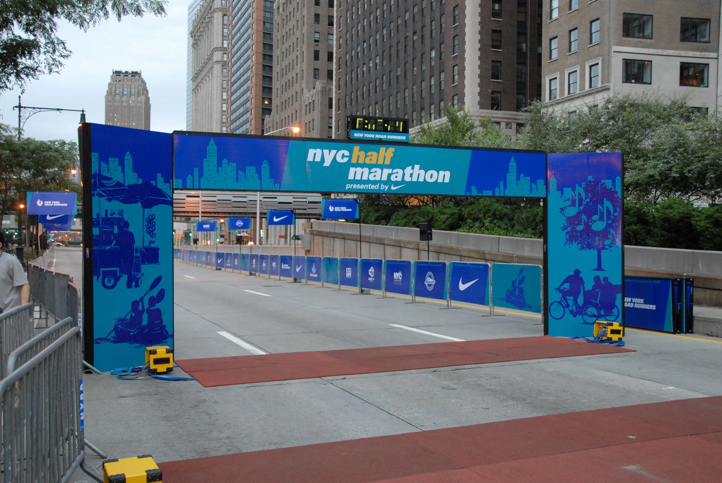 Start line for NYC Half Marathon with blue banners, tall buildings in the background, and a digital clock showing 7:04. Night is approaching in New York City.