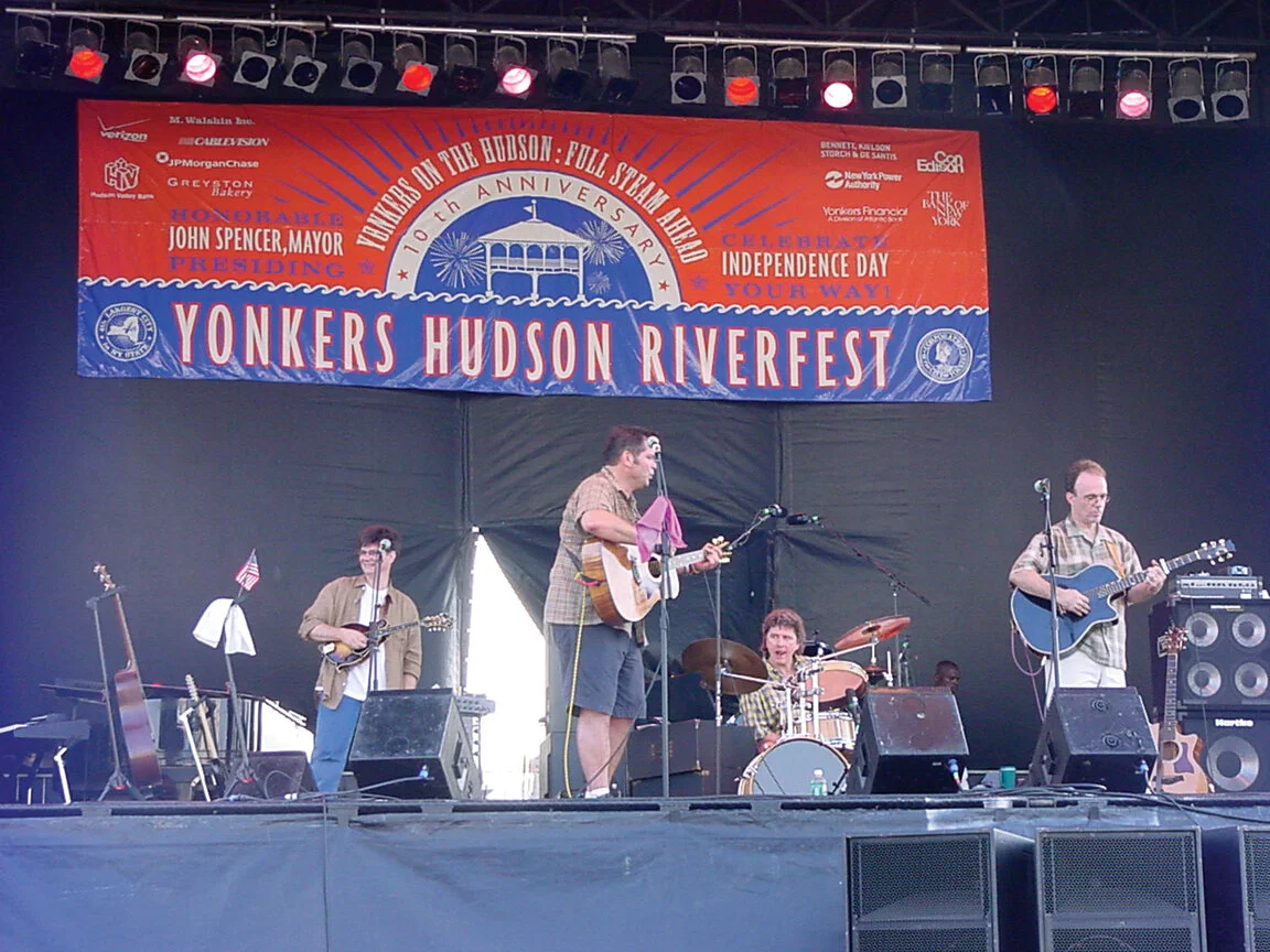 A band performing on stage at the Yonkers Hudson Riverfest, celebrating the 110th anniversary of the Hudson Full Stream Alert. The stage has a large banner with event details and red, white, and blue decorations. The band includes four members, two p