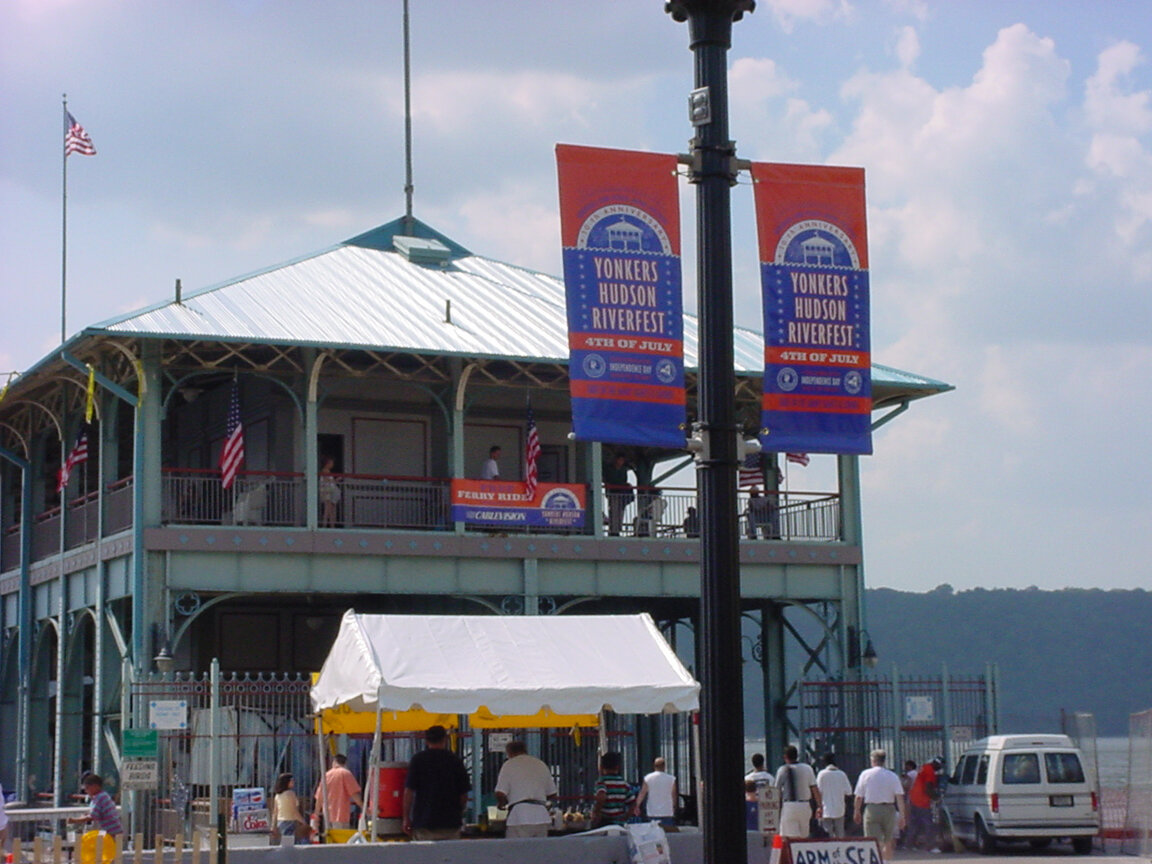 People gathering at a pier with a two-story pavilion and American flags, advertising the Yonkers, Hudson Riverfest on July 4th.