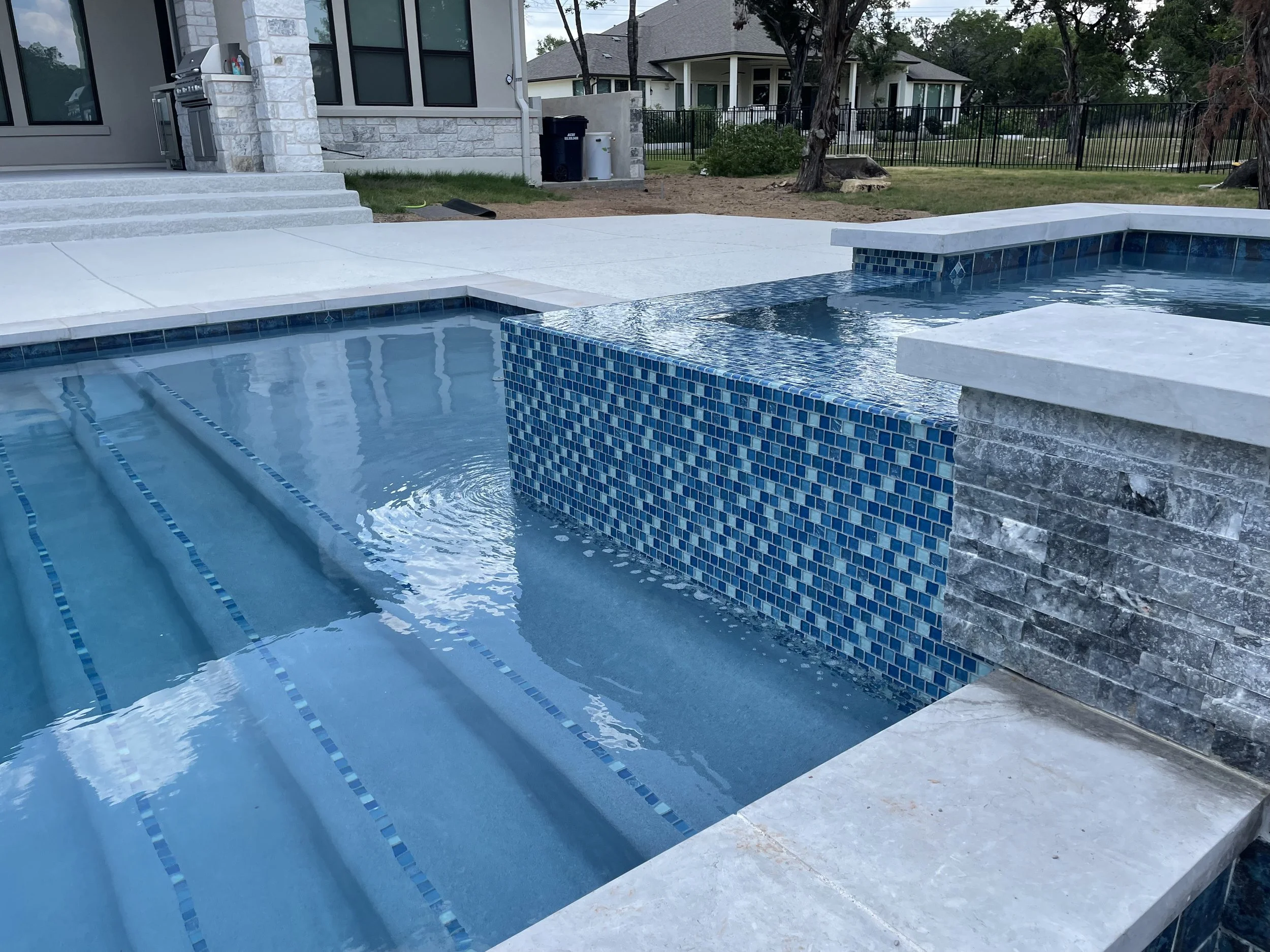 A backyard swimming pool with a built-in spa, featuring blue mosaic tile accents and a white stone deck, with a house and trees in the background.