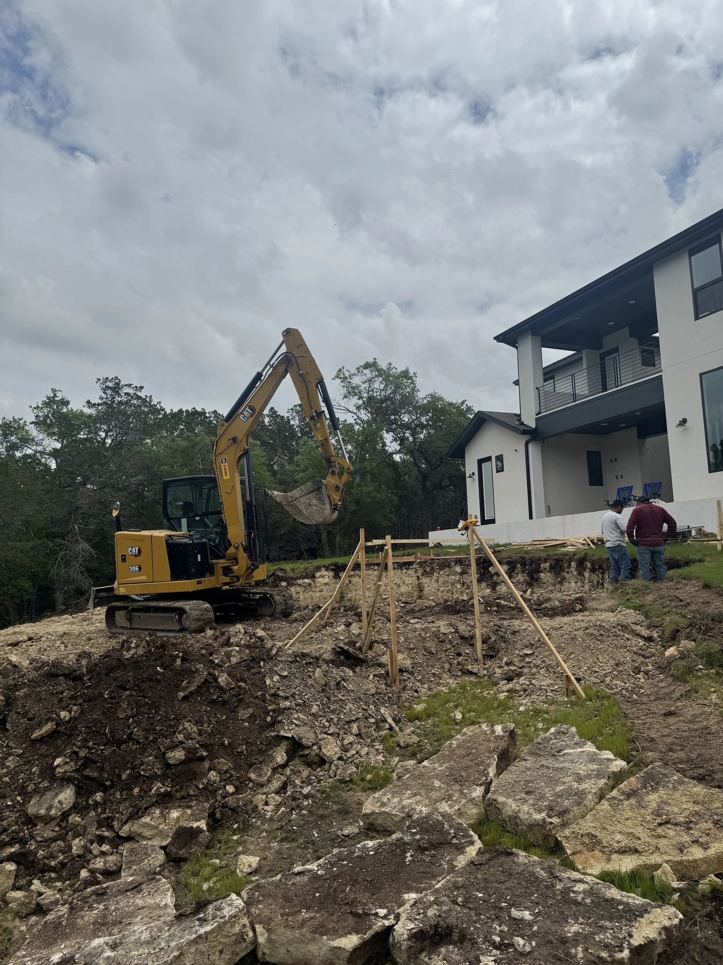 Construction site with a excavator and two workers in front of a modern house, excavation work ongoing, with large rocks and dirt in the foreground.