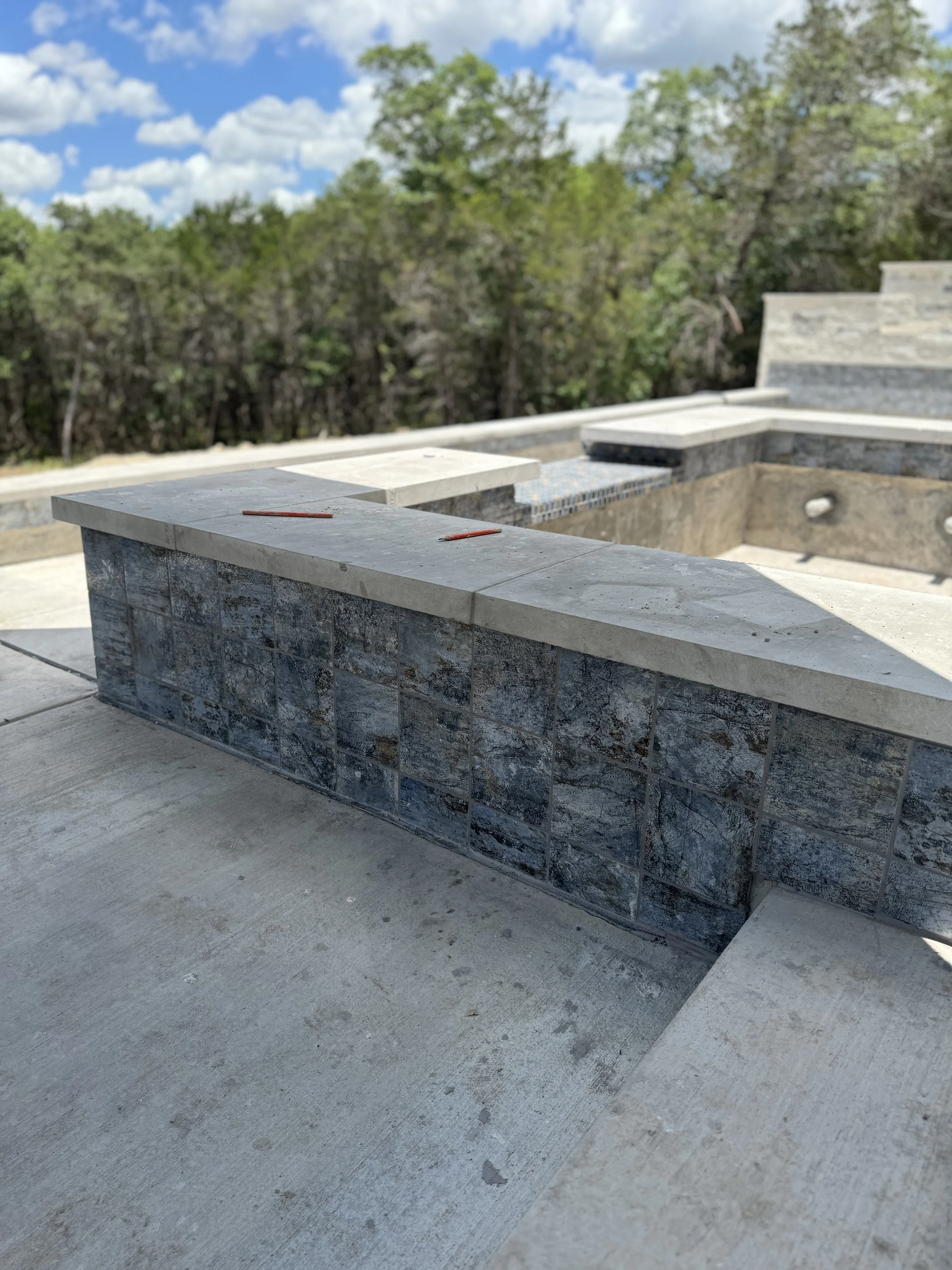 Construction site with partially built outdoor area featuring stone and concrete structures, with trees and blue sky in the background.