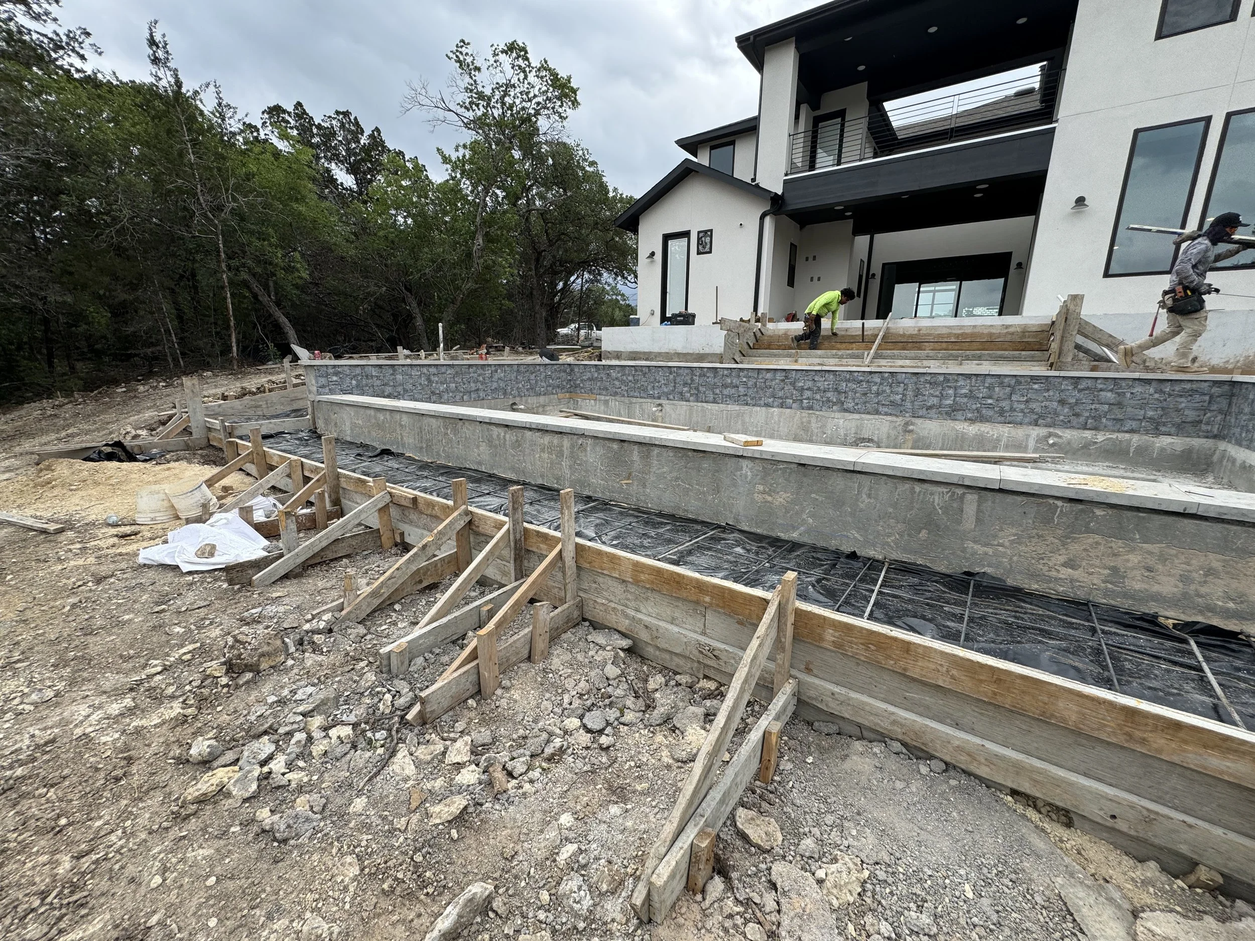 Construction workers building a large outdoor swimming pool in front of a modern white house with black accents, with steps to the house visible in the background.