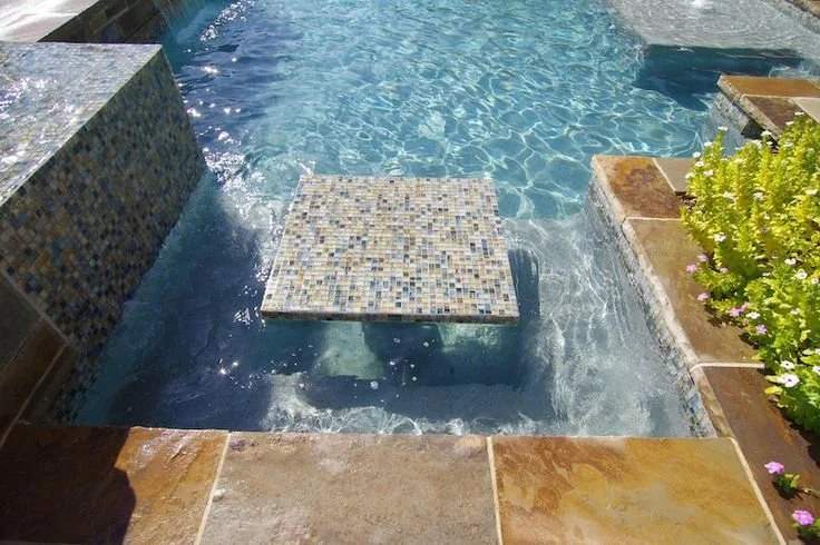 Close-up of a swimming pool with a floating mosaic-tiled platform, surrounded by stone tiles and green plants.