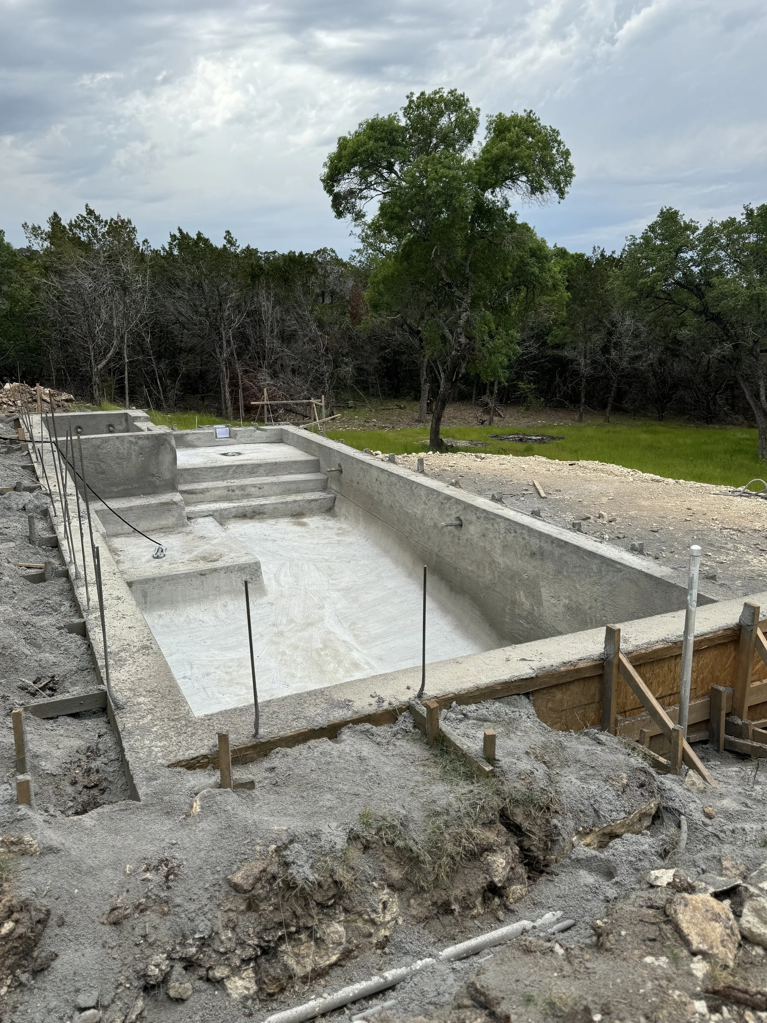 Construction site with an empty swimming pool, surrounded by dirt and construction materials, with trees and cloudy sky in the background.