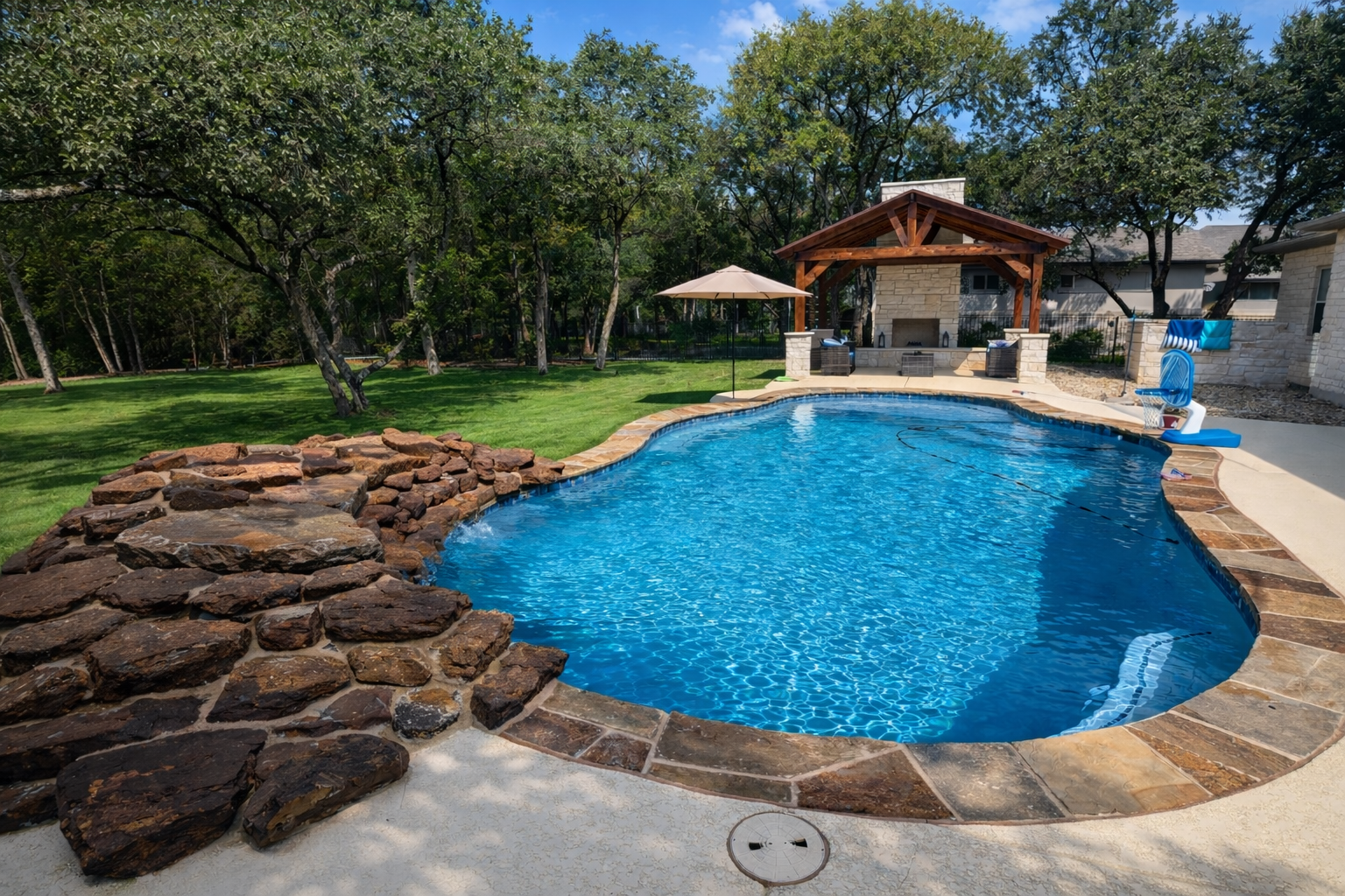 A backyard swimming pool with a stone border, surrounded by green grass and trees, featuring a shaded outdoor area with a roof, chairs, and a large umbrella. There is a blue sign with five gold stars and the words "5 Star Rated Pool."