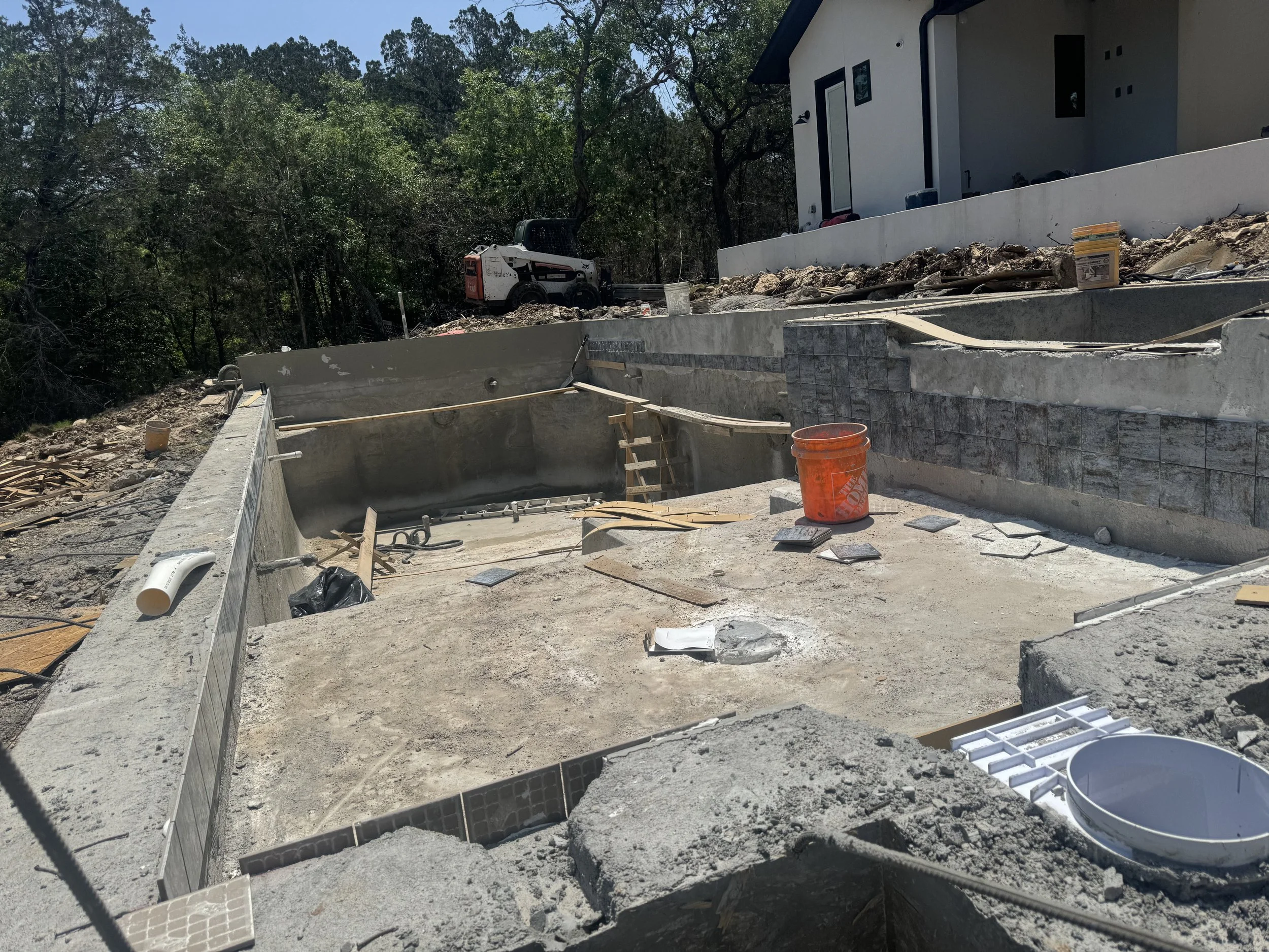 Under construction in-ground swimming pool area with unfinished concrete surfaces, construction tools, and materials, with a house and wooded background.