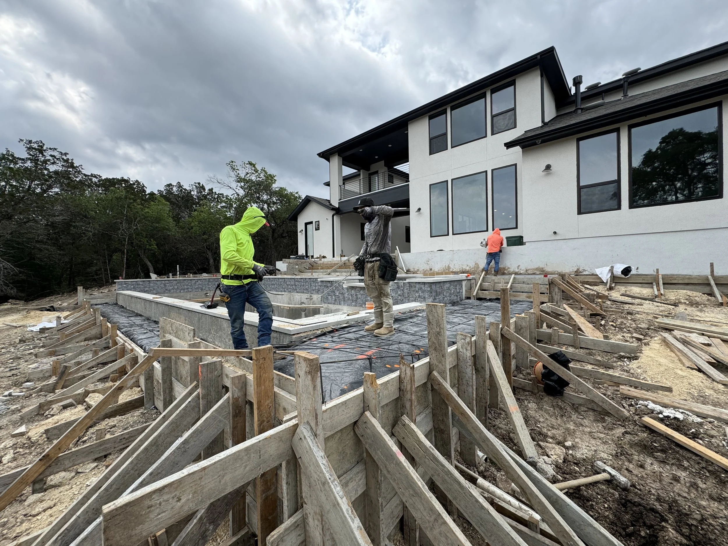 Construction workers pouring and leveling concrete for a patio or pool area in front of a modern house during daytime with cloudy sky.