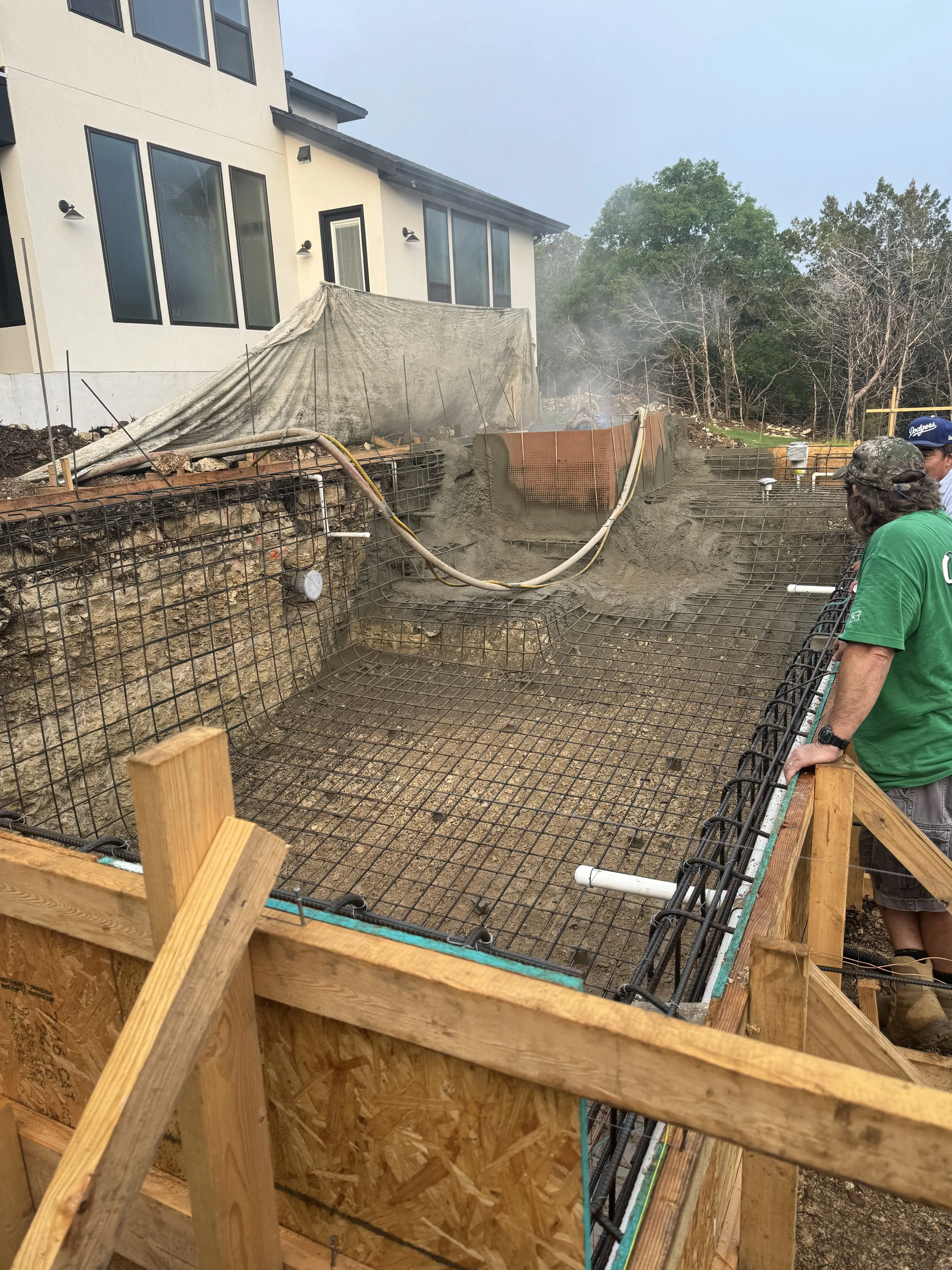 Construction site with a house in the background, showing foundation work for a swimming pool with rebar and concrete, and people overseeing the process.