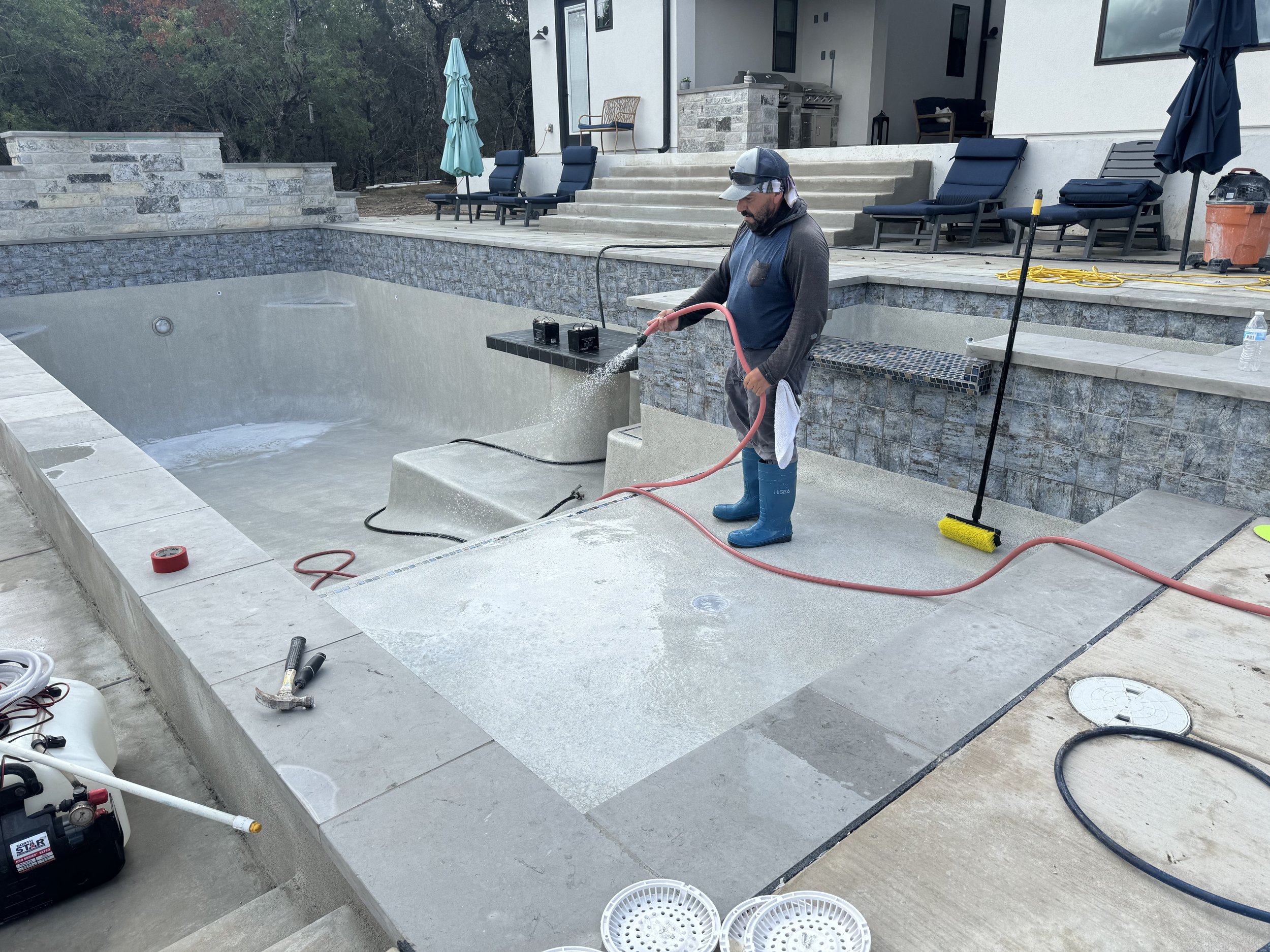 A man in blue boots and a cap is cleaning a filled-in swimming pool with a garden hose. The pool is surrounded by a stone patio, and there are lounge chairs, umbrellas, and a house in the background.