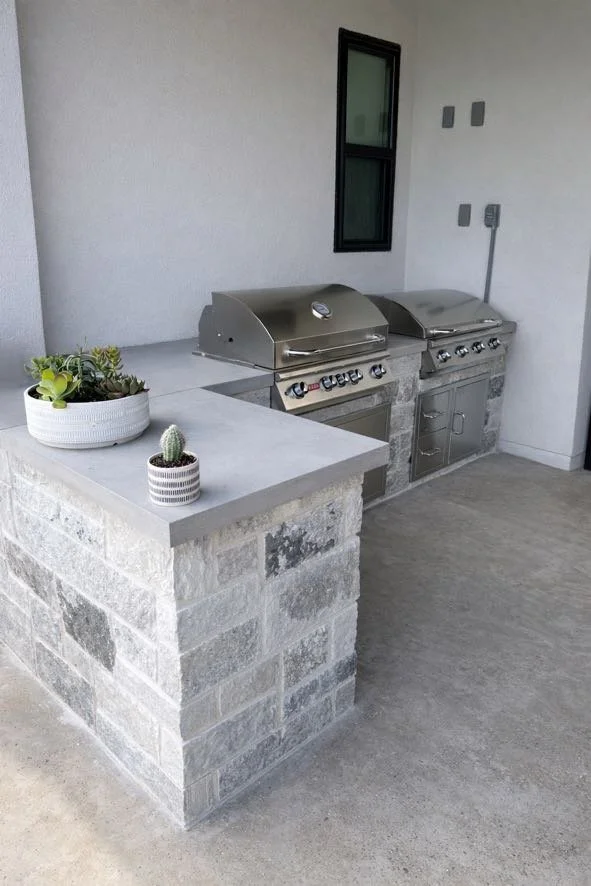 Outdoor kitchen area with two stainless steel gas grills on a stone counter, a small potted cactus and a plant in a white pot on the side, a small window above, and a gray wall with electrical outlets.