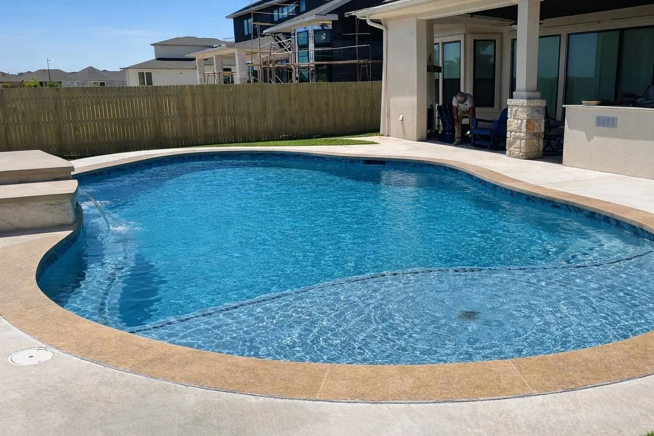 Backyard swimming pool with spa area and a covered patio, a man sitting on a chair, neighboring houses, wooden fence, green grass, clear blue sky.