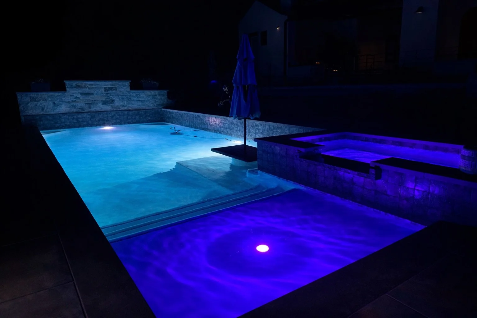 Illuminated swimming pool at night with blue and purple LED lights, an umbrella, and a hot tub nearby.