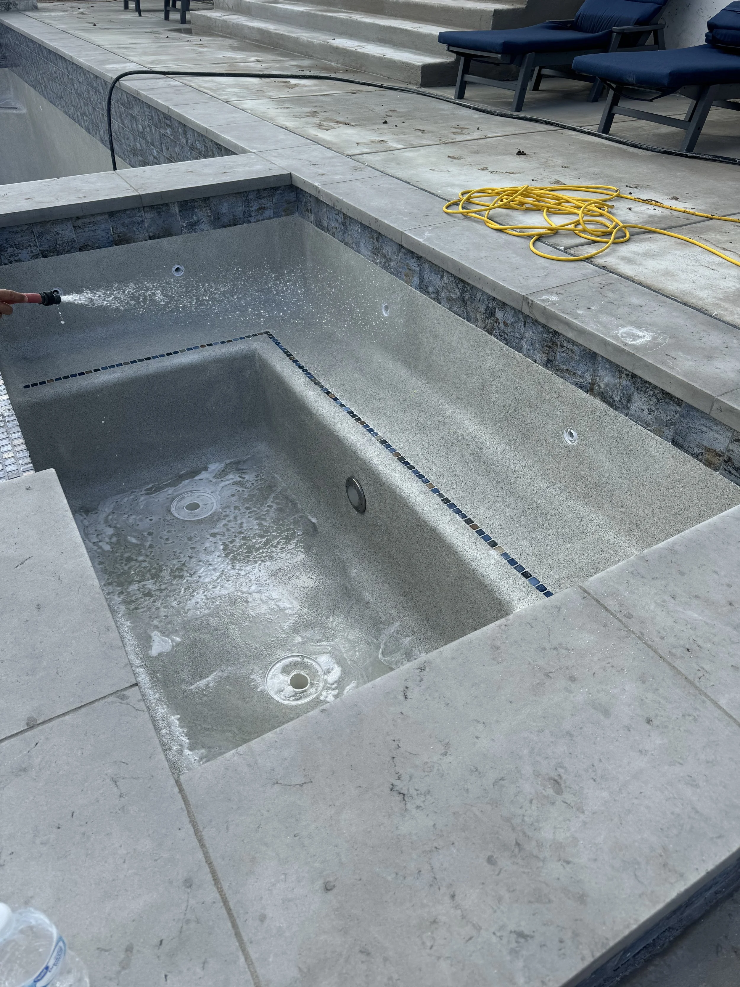 Empty swimming pool being cleaned with a hose. The pool has light-colored interior with small decorative tiles and is surrounded by a concrete deck. There are blue chairs and a yellow extension cord on the patio nearby.
