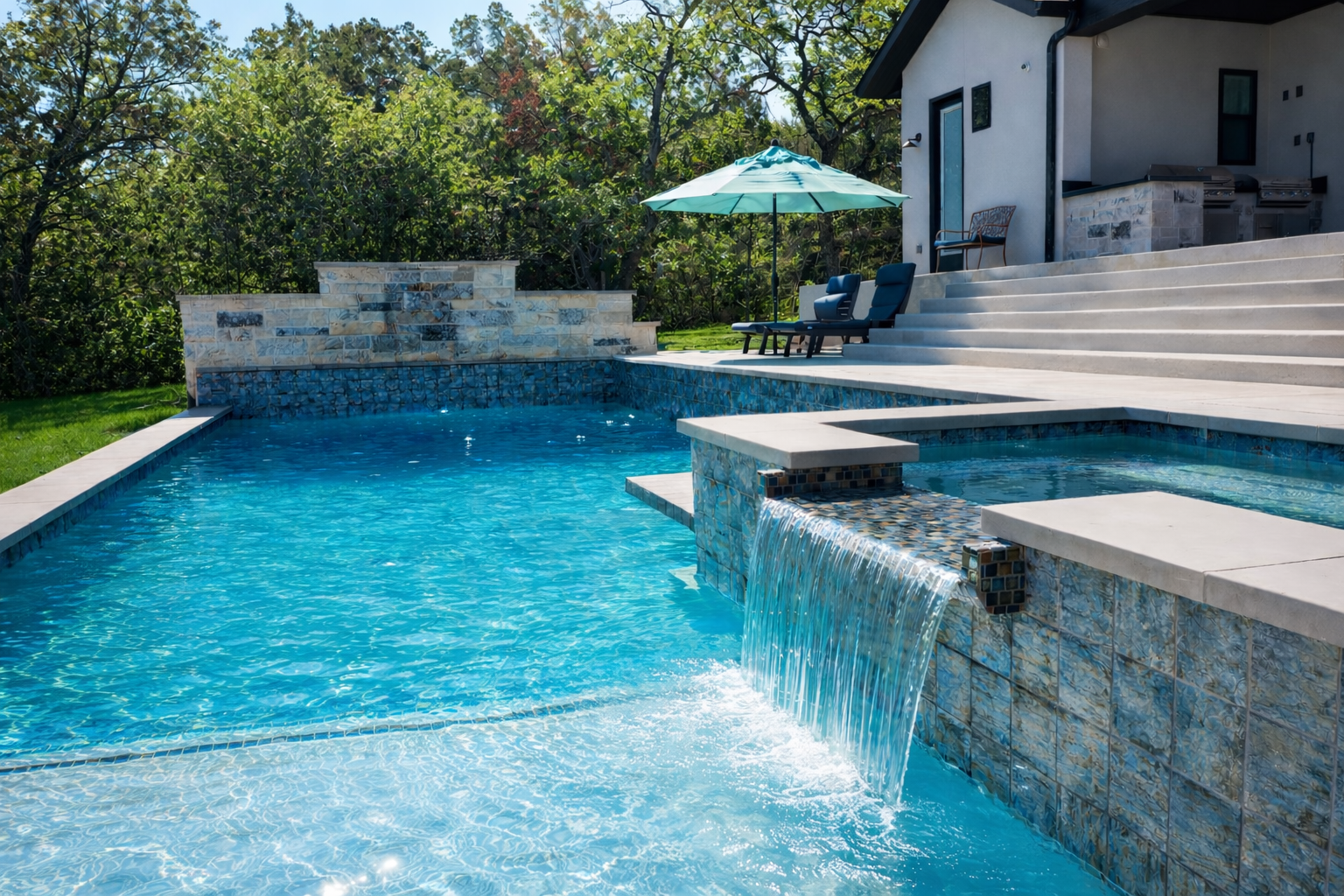Backyard swimming pool with a waterfall feature, poolside lounge chairs with cushions, sun umbrella, in-ground hot tub, and a house with outdoor cooking area, surrounded by trees.
